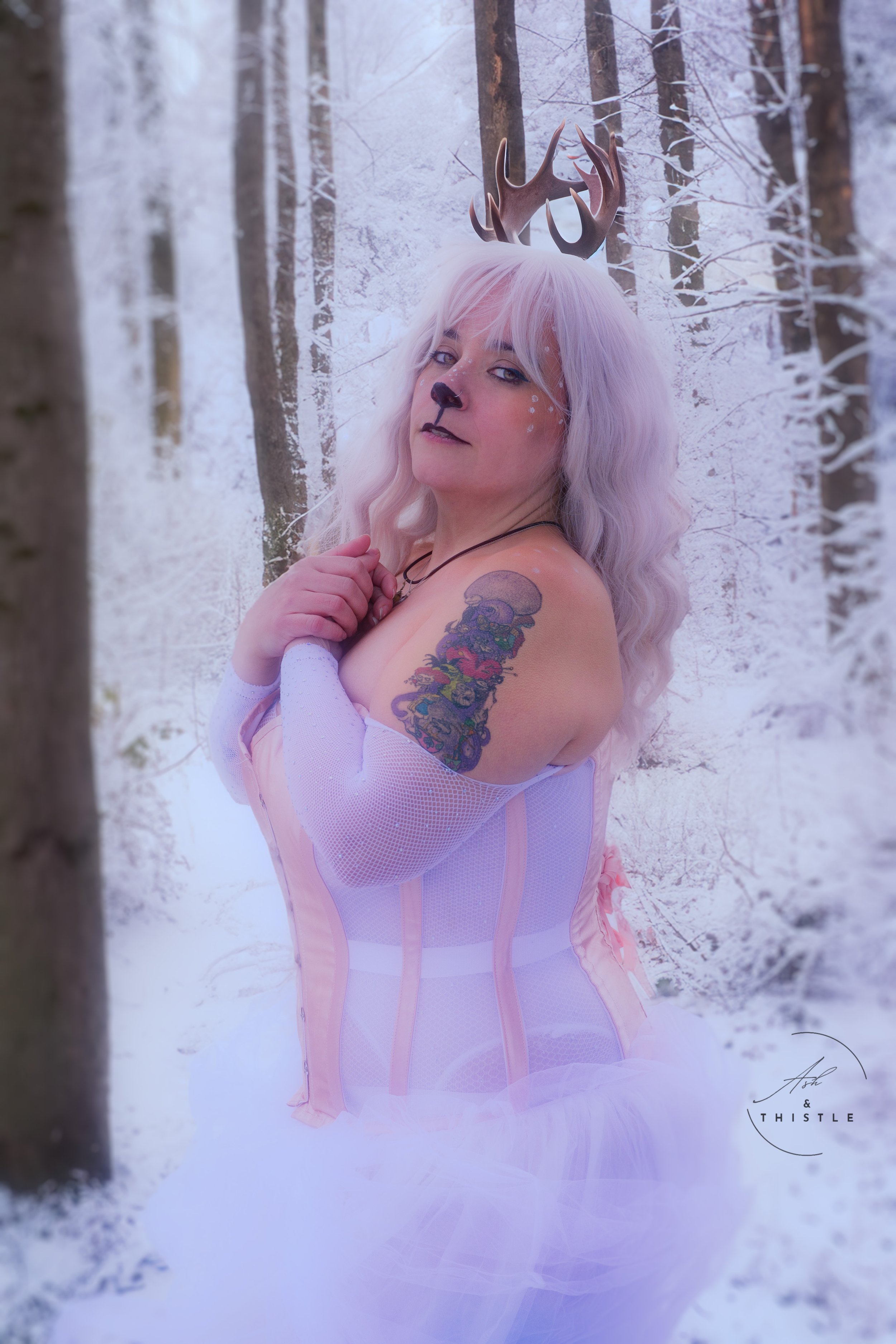 Person dressed in deer costume with antlers, white wig, and makeup in snowy forest.