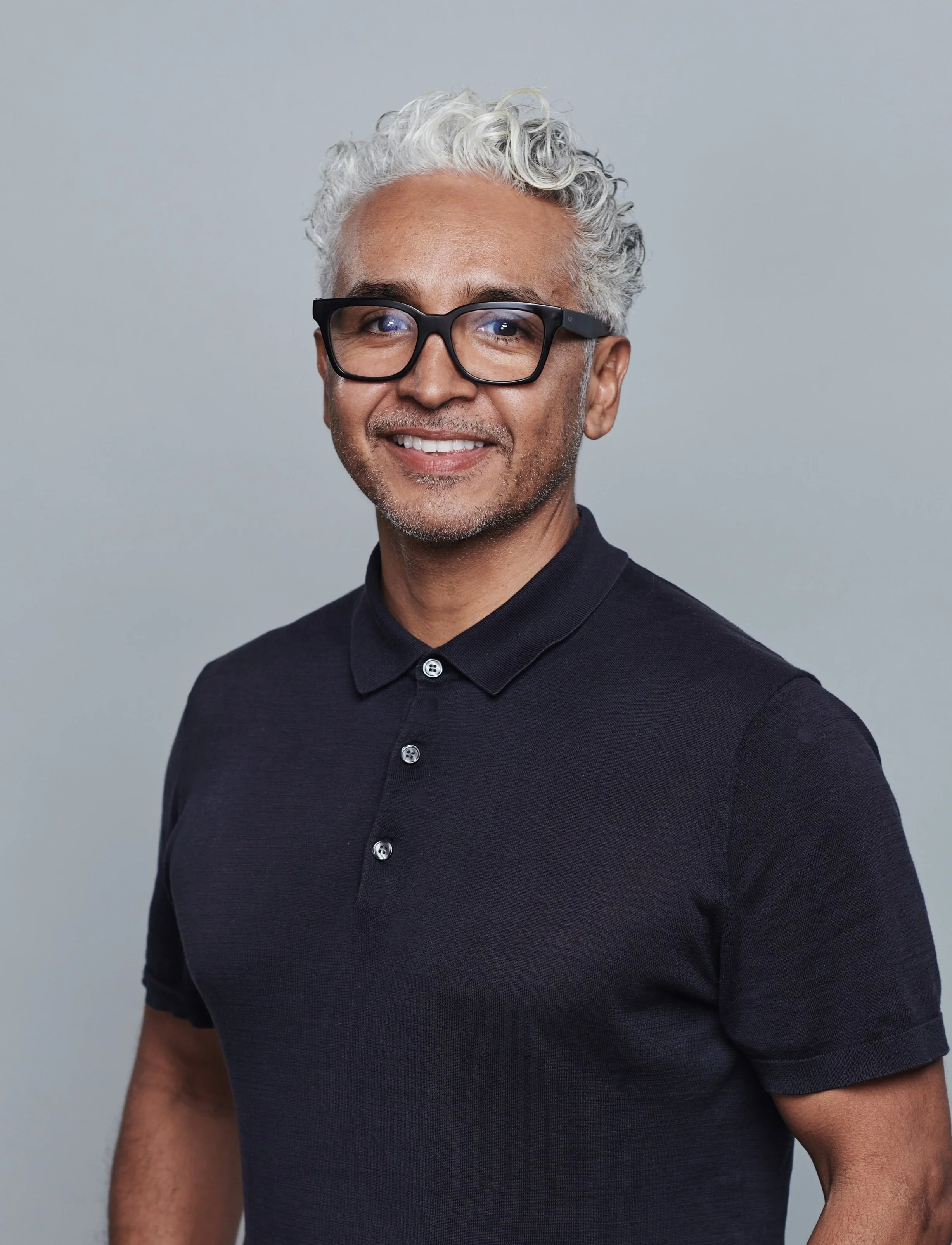 A middle-aged man with curly gray hair and black glasses, wearing a dark polo shirt, smiling at the camera against a neutral background.