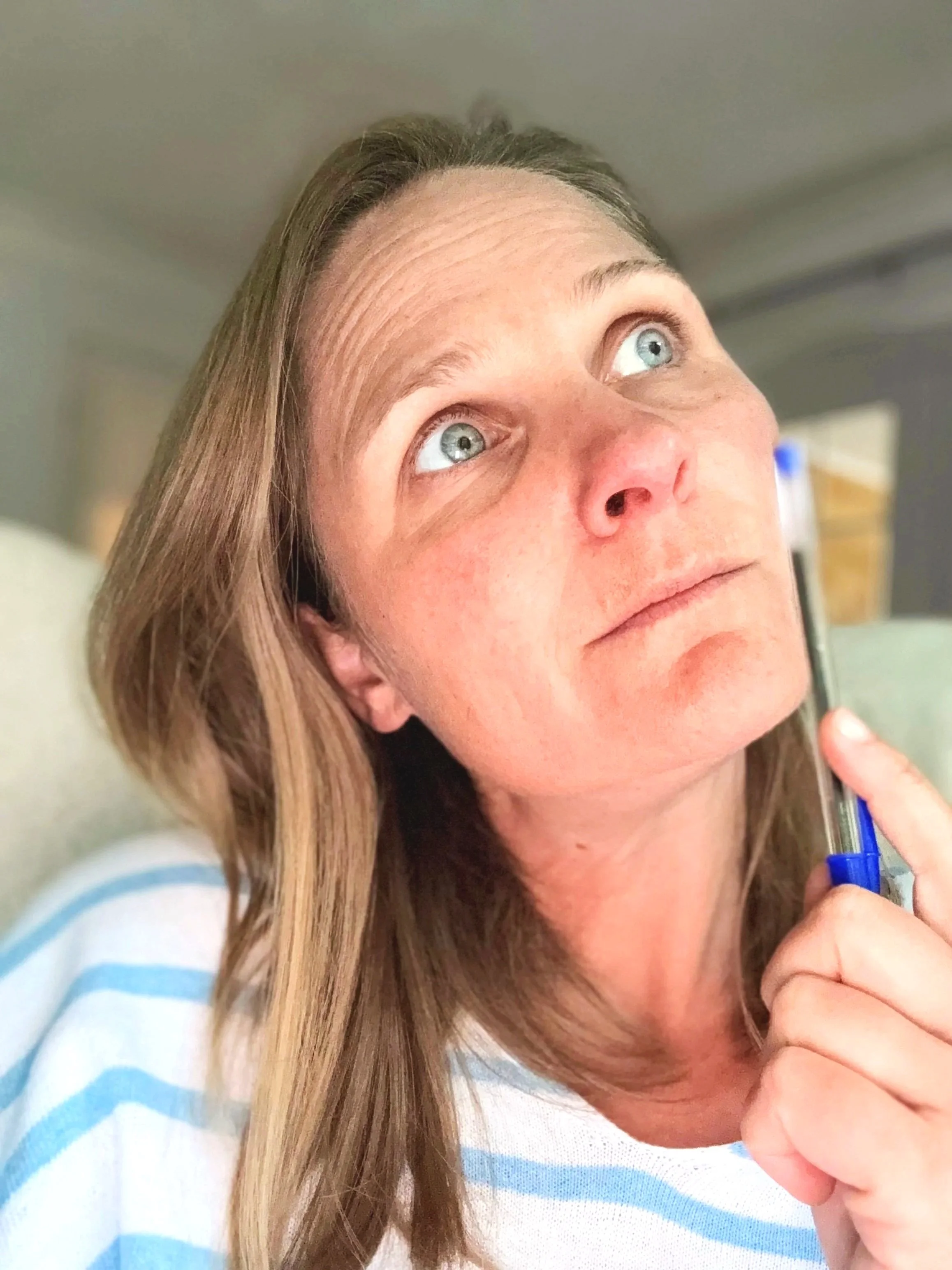 A woman with long blonde hair looking up and to the side, holding a pen near her chin, wearing a blue and white striped shirt, indoors.