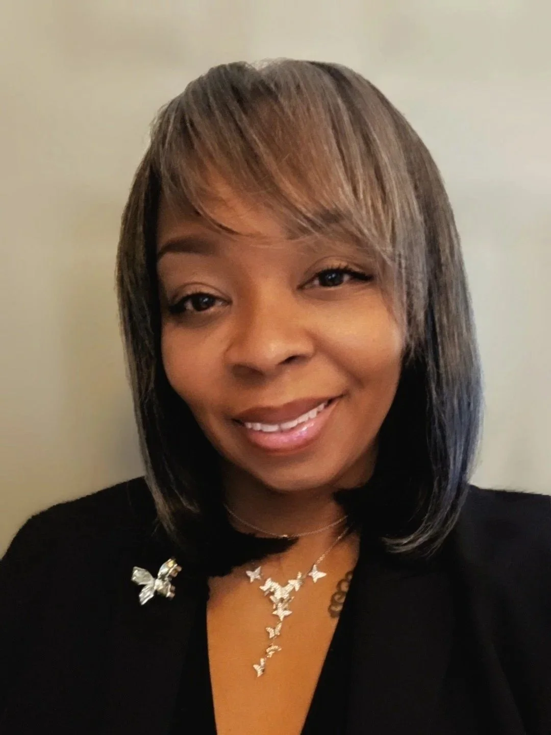 A woman with shoulder-length hair styled with highlights, wearing a black top and a silver butterfly brooch. She has on a necklace with butterfly pendants and is smiling.