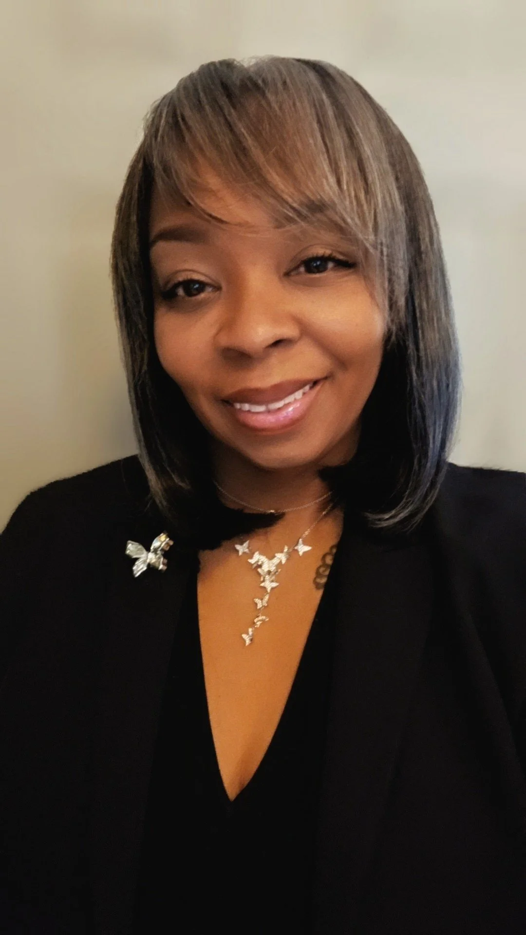 Smiling woman with shoulder-length hair, wearing a black top and butterfly-shaped jewelry, standing against a plain wall.