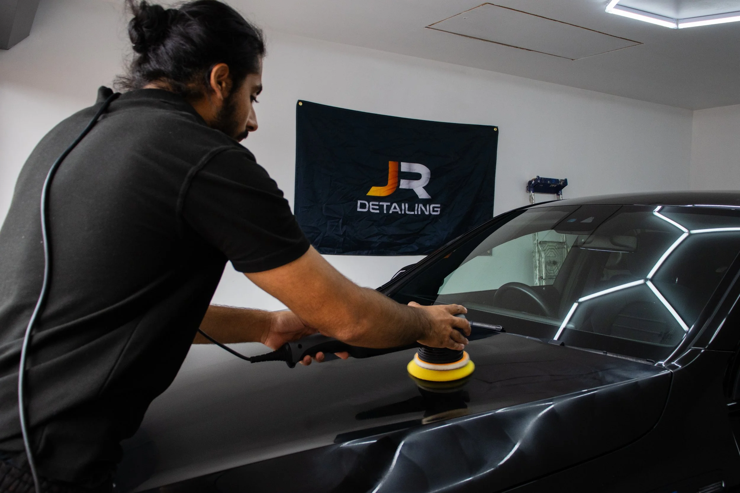 A person polishing the exterior of a black car inside a garage with a banner on the wall behind them displaying 'JR Detailing'.