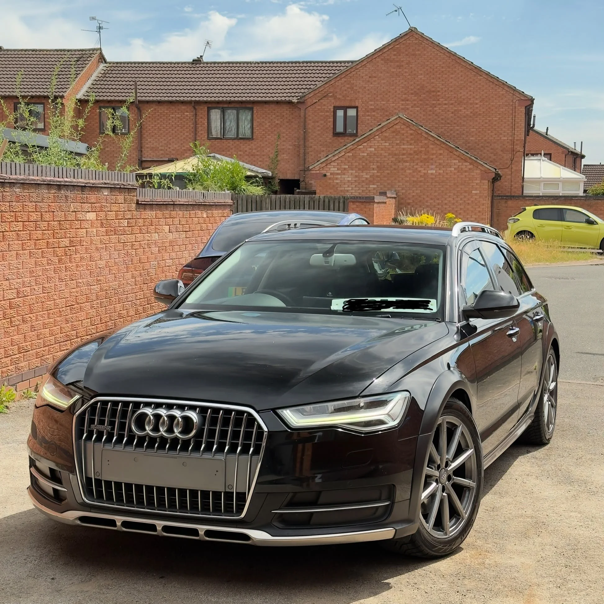Black Audi car parked in front of a brick wall and houses with pitched roofs in a residential area.