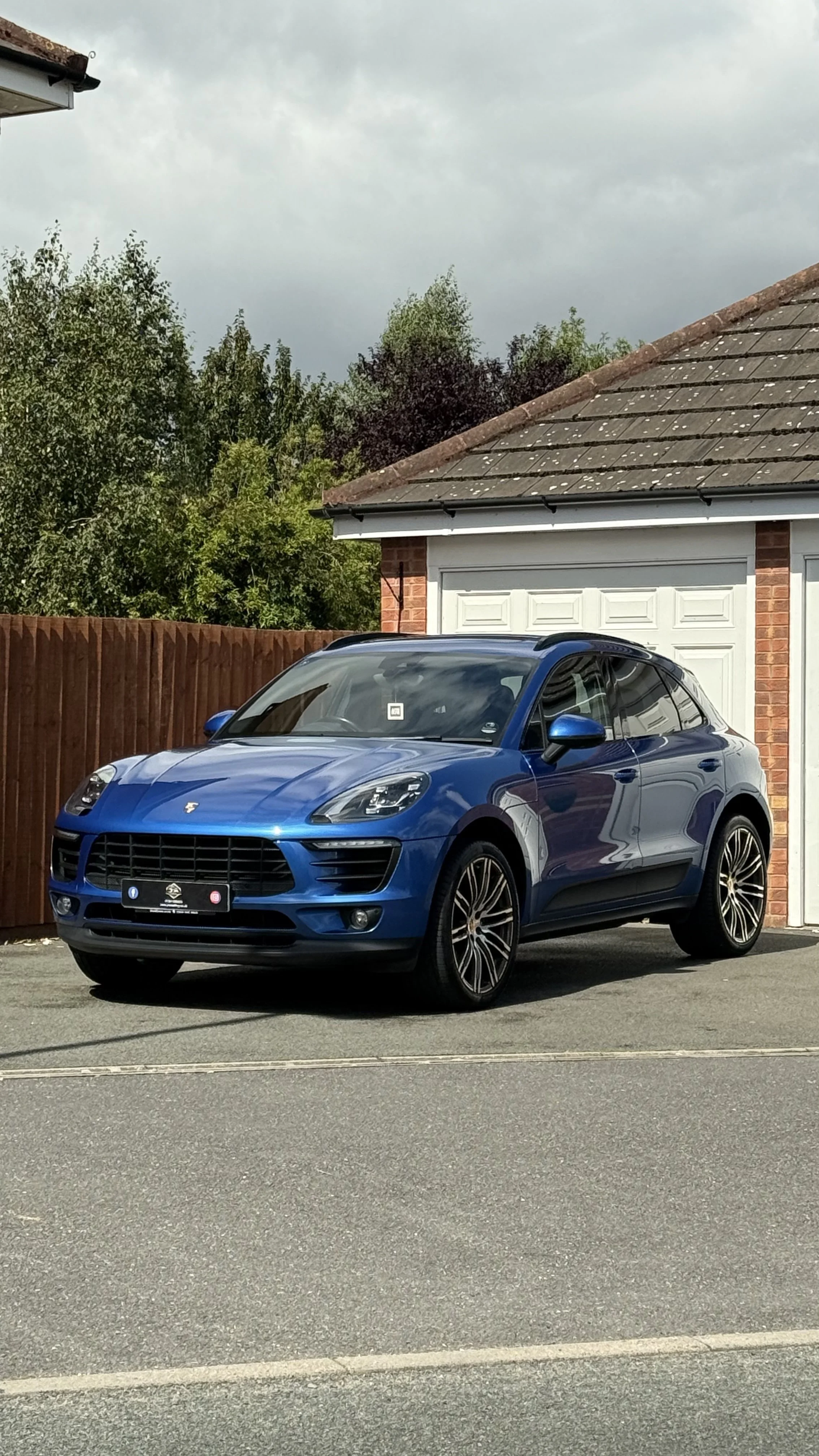 Blue Porsche Macan parked in front of a garage with a brick house and wooden fence in the background.