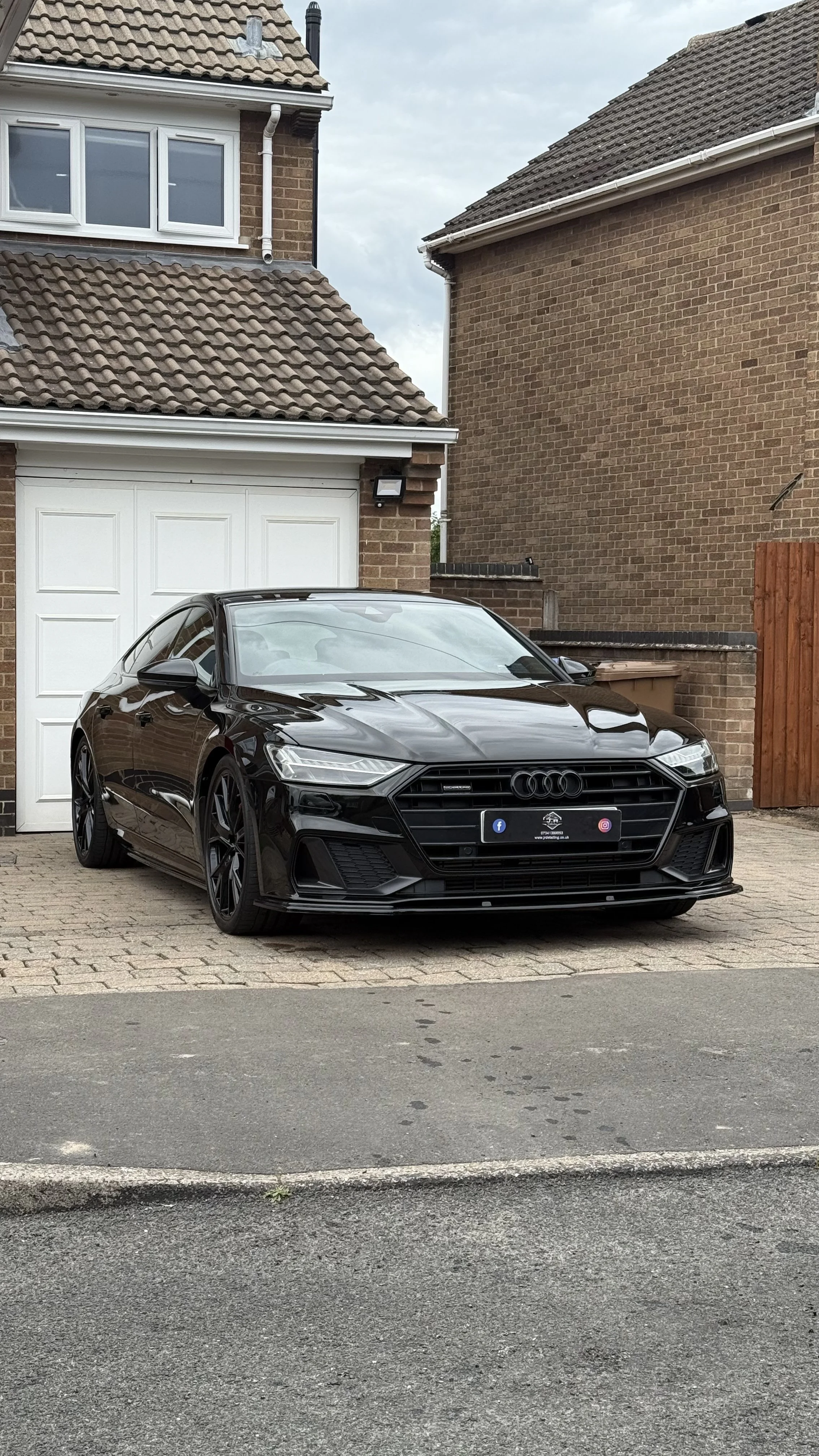 Black Audi sedan parked in front of a suburban house with a white garage door and brick exterior.