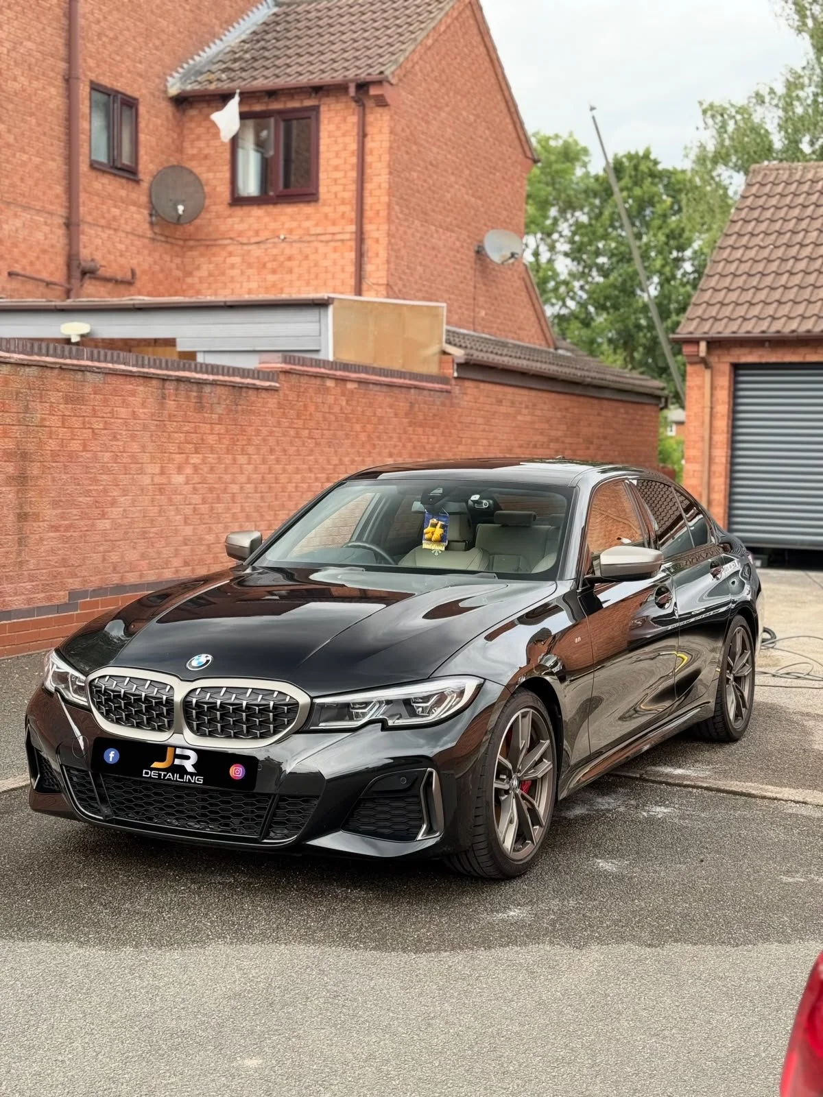 A black BMW car parked in front of a brick house with a tiled roof, satellite dishes, and a backyard fence. The car has a detailed front grille and modern headlights.