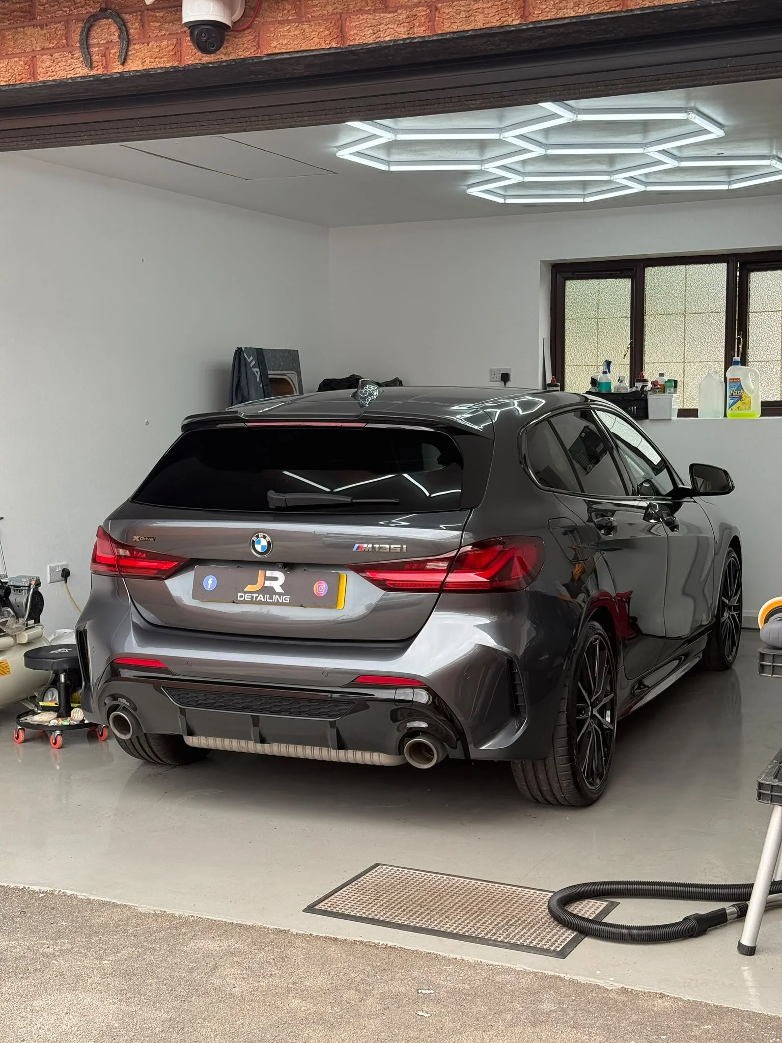 A grey BMW M135i parked inside a garage. The garage has white walls, a window with laundry detergent bottles and cleaning supplies on the ledge, and a modern hexagonal ceiling light. There are cleaning tools and appliances on the floor and a shelving unit in the background.