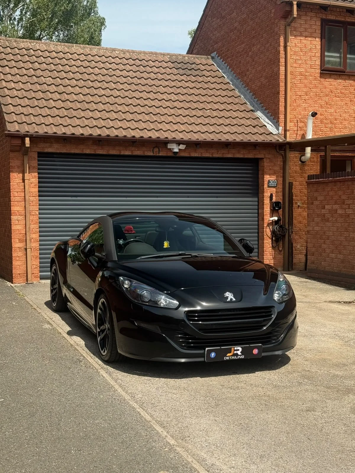 A black Peugeot convertible parked in front of a brick garage with a closed metal shutter door. The house has a red brick exterior and a tiled sloped roof with greenery visible in the background.