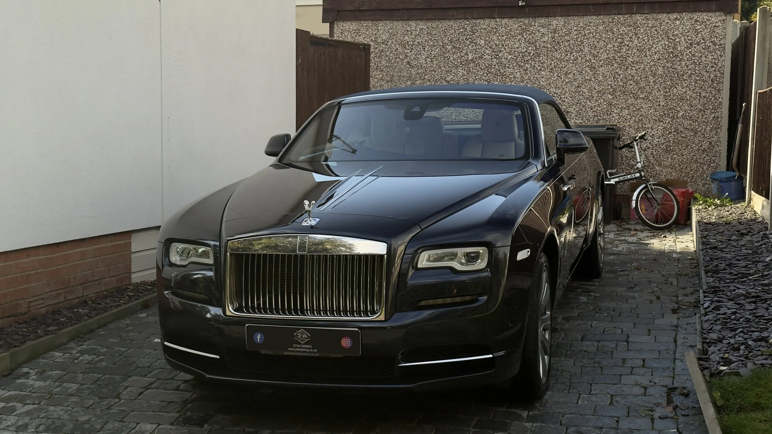 A black Rolls-Royce luxury car parked on a cobblestone driveway next to a bicycle, with a white wall, brick wall, and wooden fence in the background.