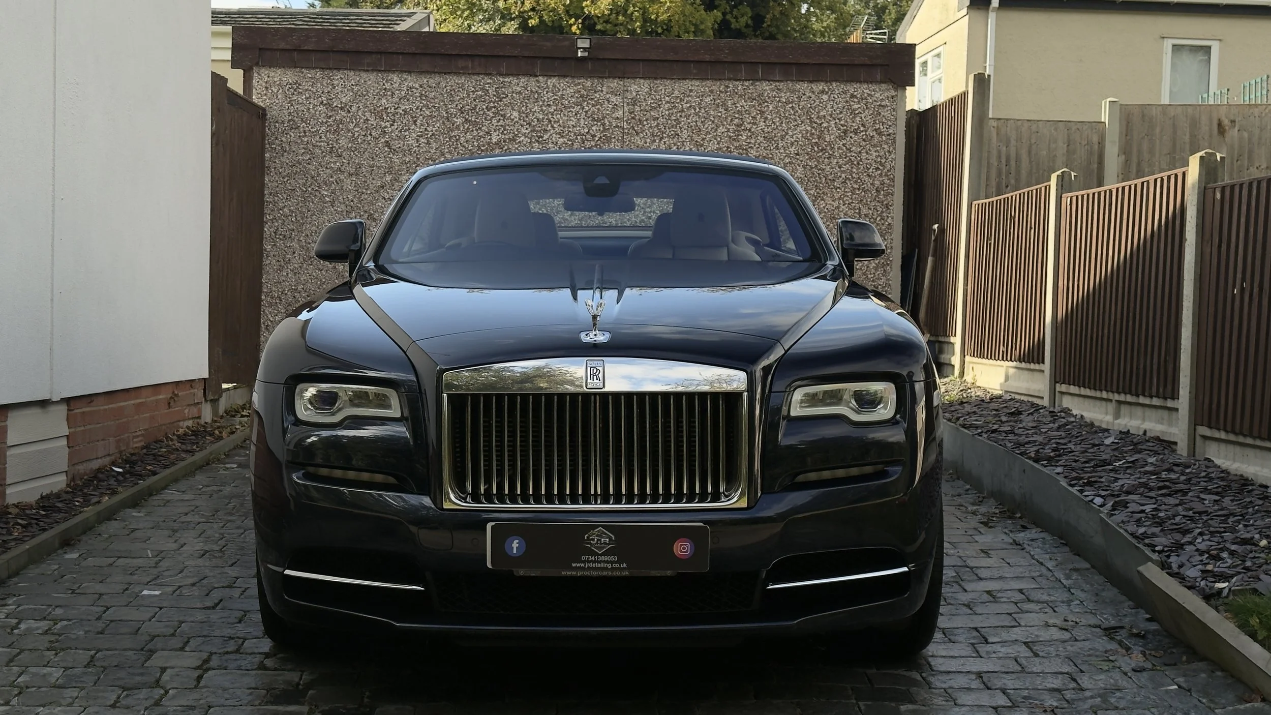 A black Rolls-Royce car parked on a brick driveway, flanked by wooden fences and residential buildings.