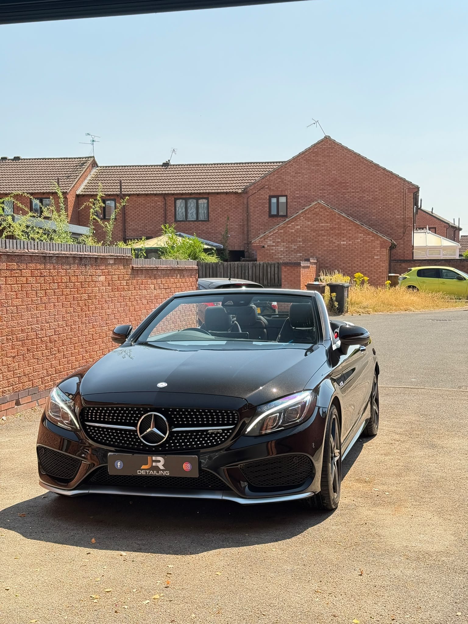 A black Mercedes-Benz convertible car parked in a residential area with brick houses and a clear blue sky in the background.