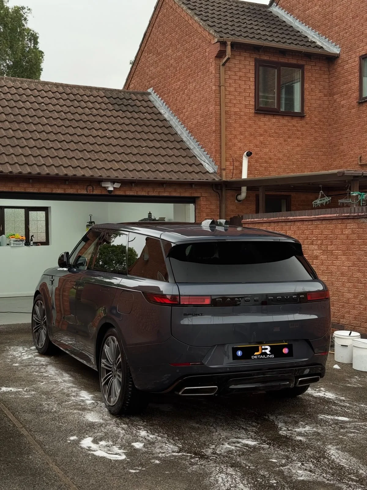 A black Range Rover Sport parked in a driveway next to a brick house. The driveway has some snow and ice. The house has brown tiled roofs, windows, and accessories, with a small porch area.