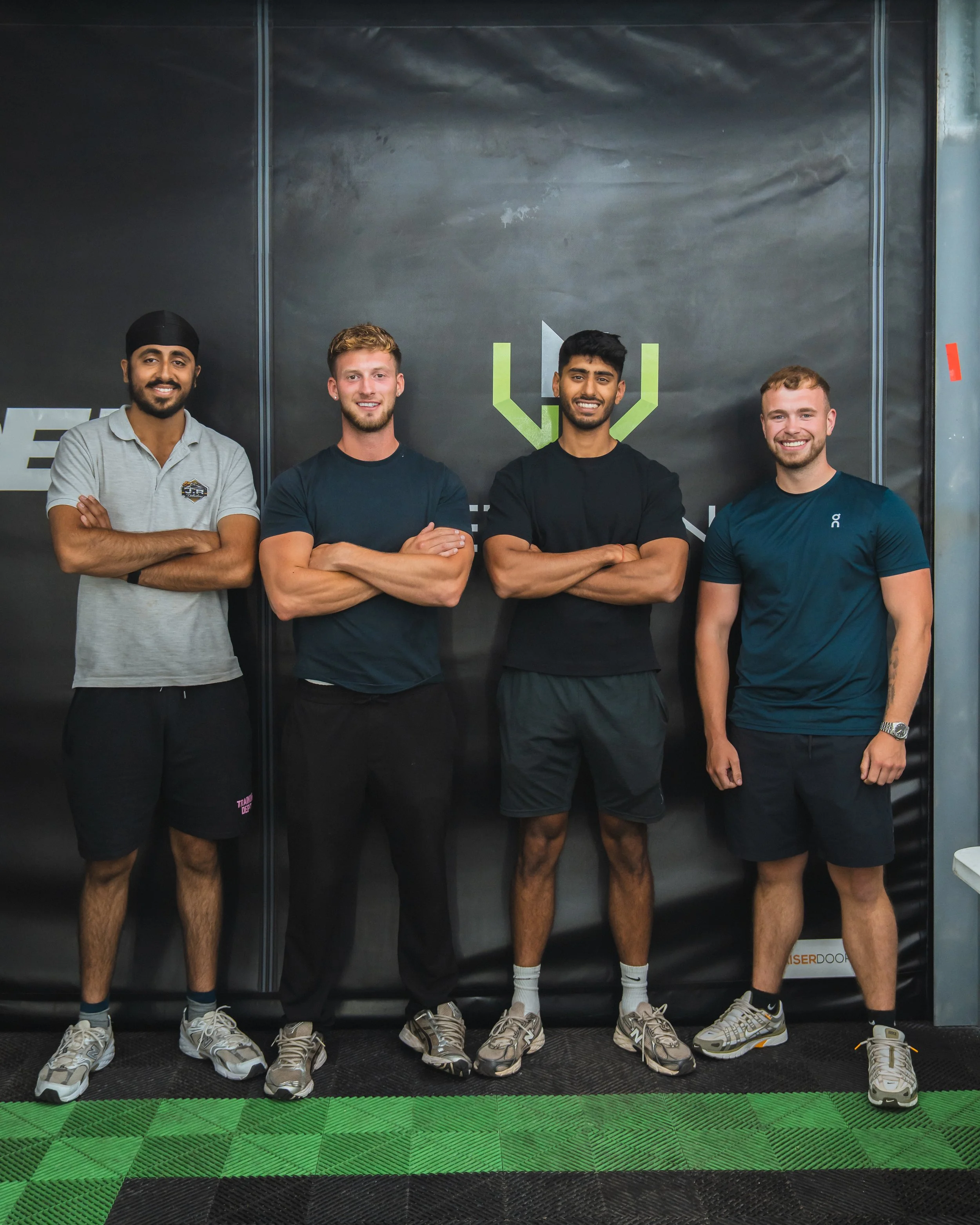 Four men standing with arms crossed, smiling, in front of a black wall with a green and white logo, in a gym or training room.