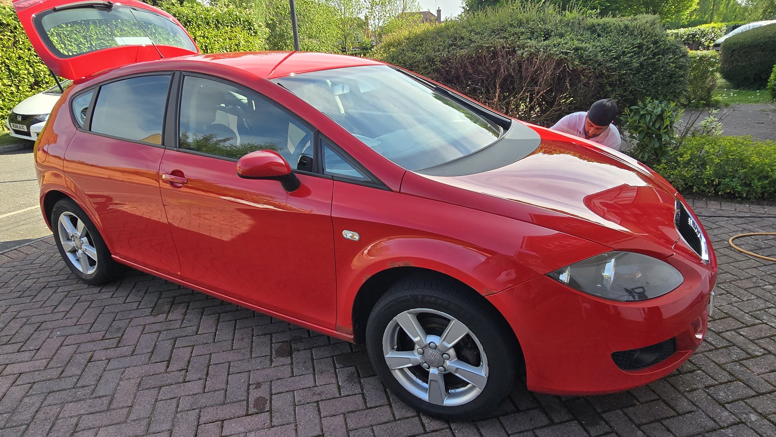 Red hatchback car parked on driveway with open trunk, person cleaning nearby, green hedges in background.