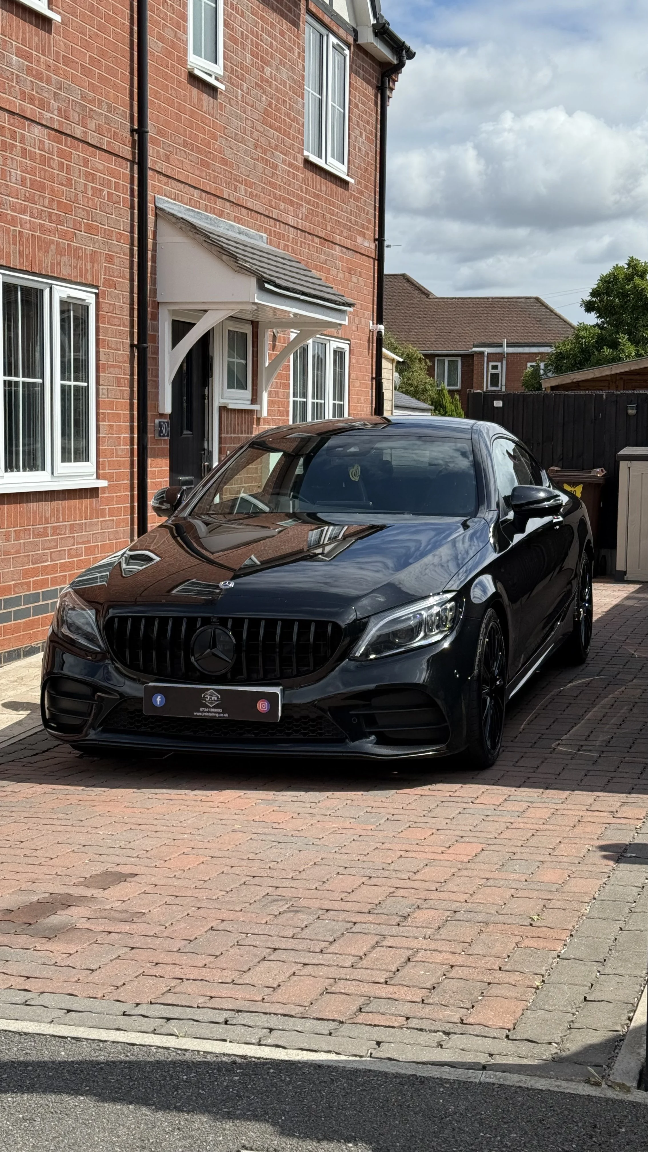 Black Mercedes-Benz car parked in front of a red brick house with white window frames on a paved driveway.