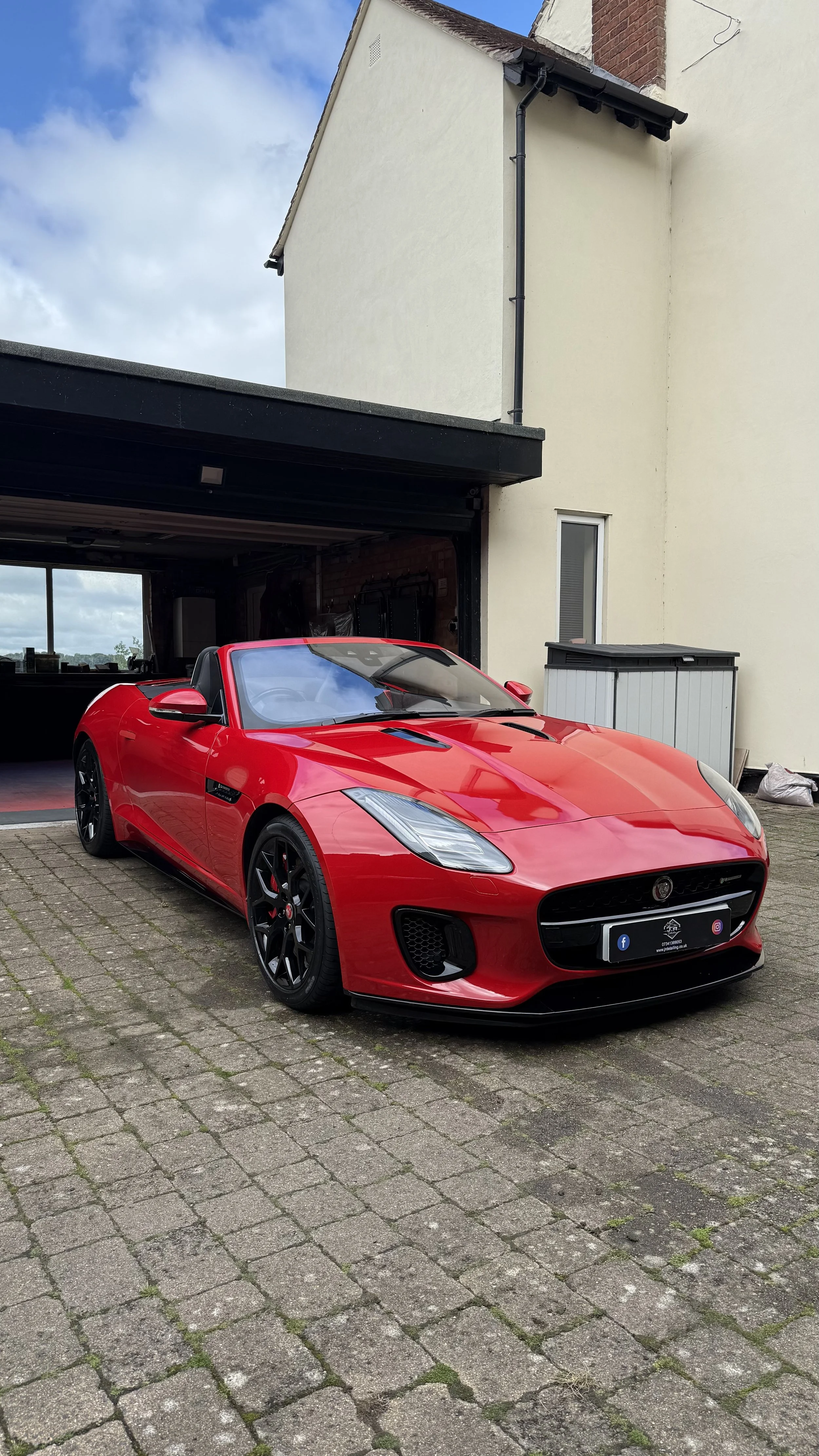 A red convertible sports car parked on a brick driveway outside a house with a white exterior and black roof.