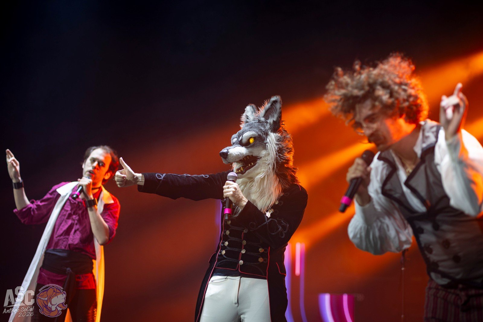 Pom, Chrono, and Charlie on stage, singing into microphones. Chrono is pointing toward the audience, with colorful red and orange stage lighting.