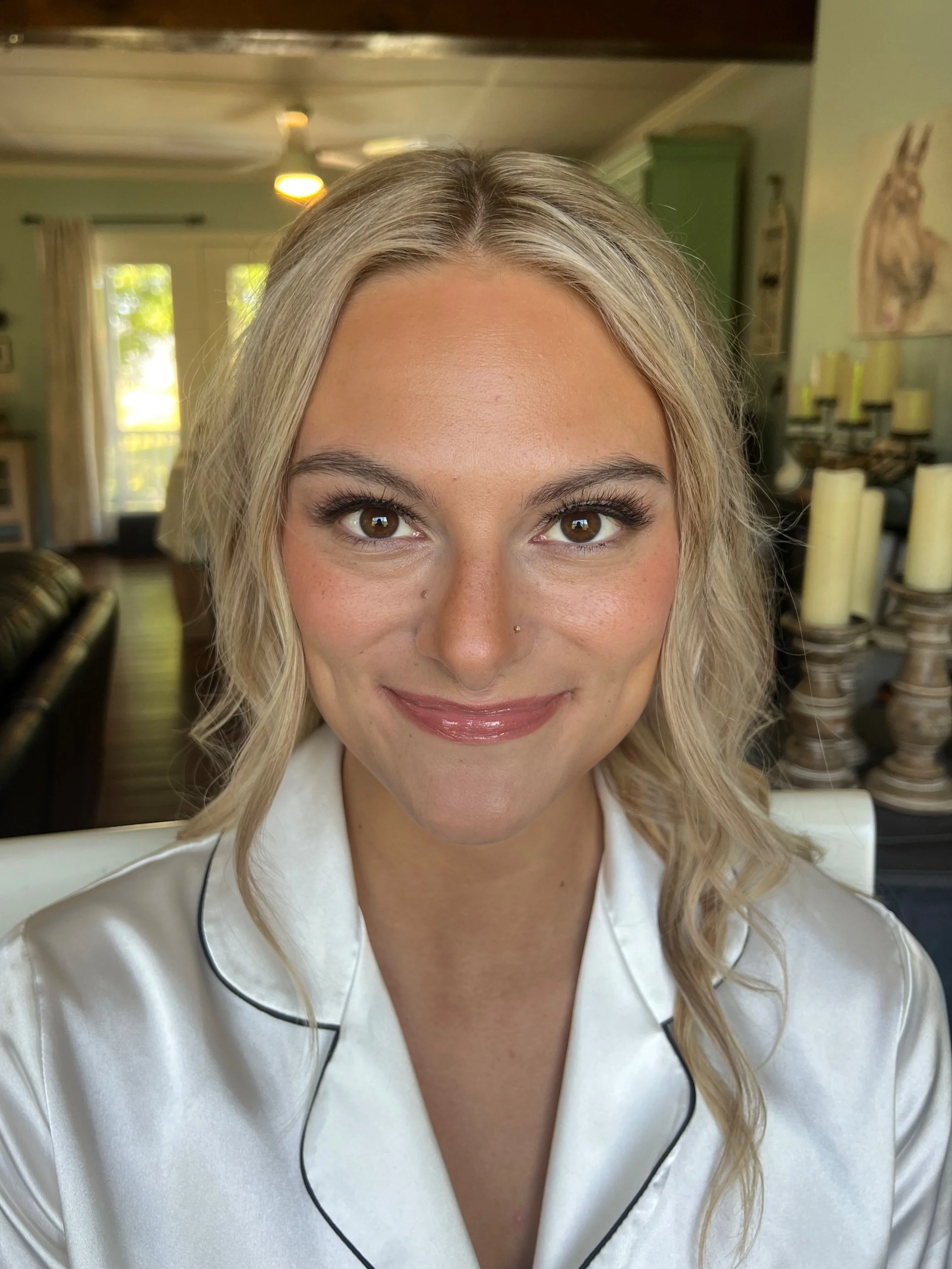 A woman with blonde wavy hair, wearing a cream-colored satin pajama top with black piping, smiling in a well-lit room with green walls, candles, and a window in the background.