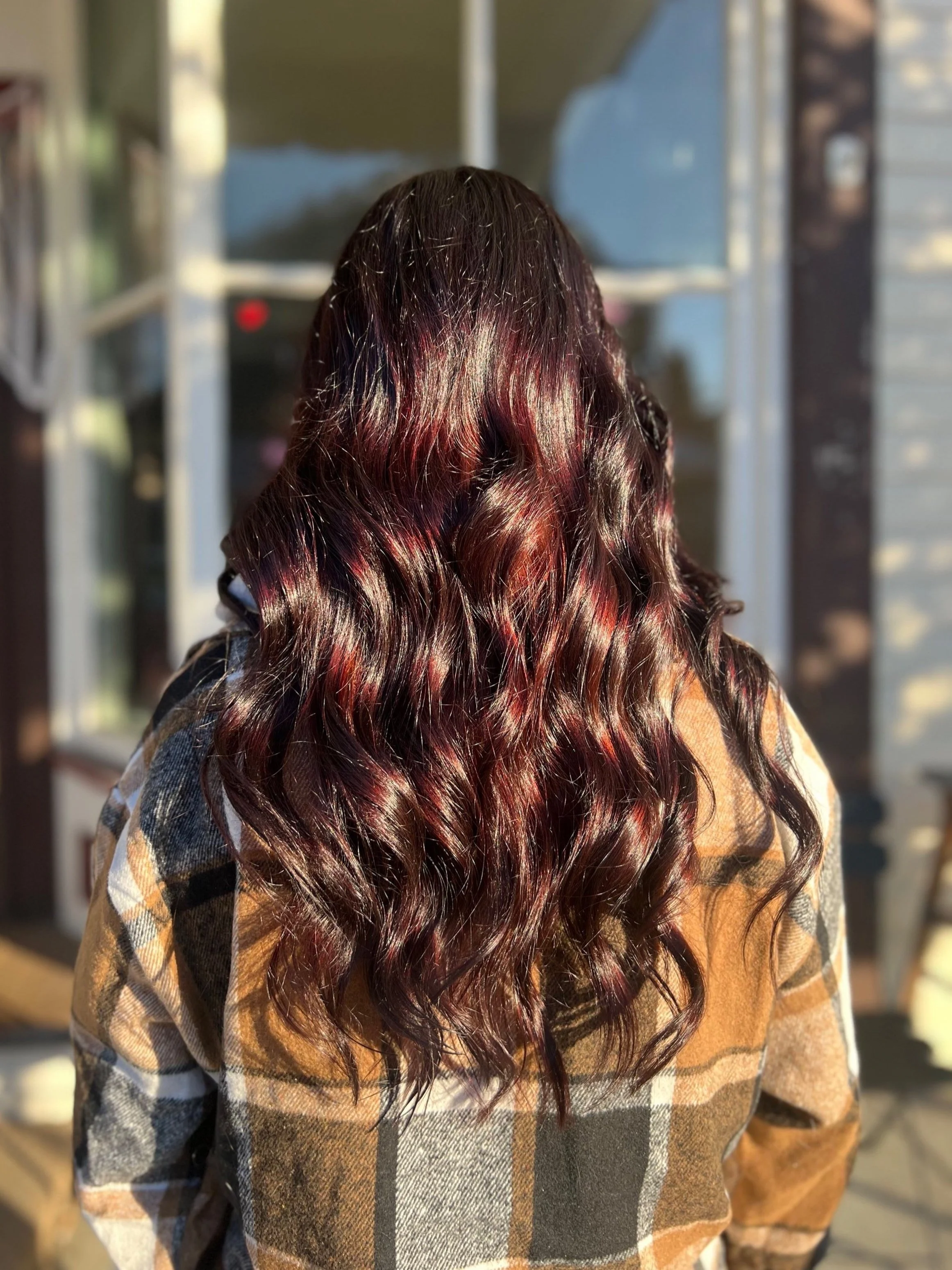 Back view of a person with long, curly, burgundy hair, wearing a plaid shirt, standing outdoors in sunlight.