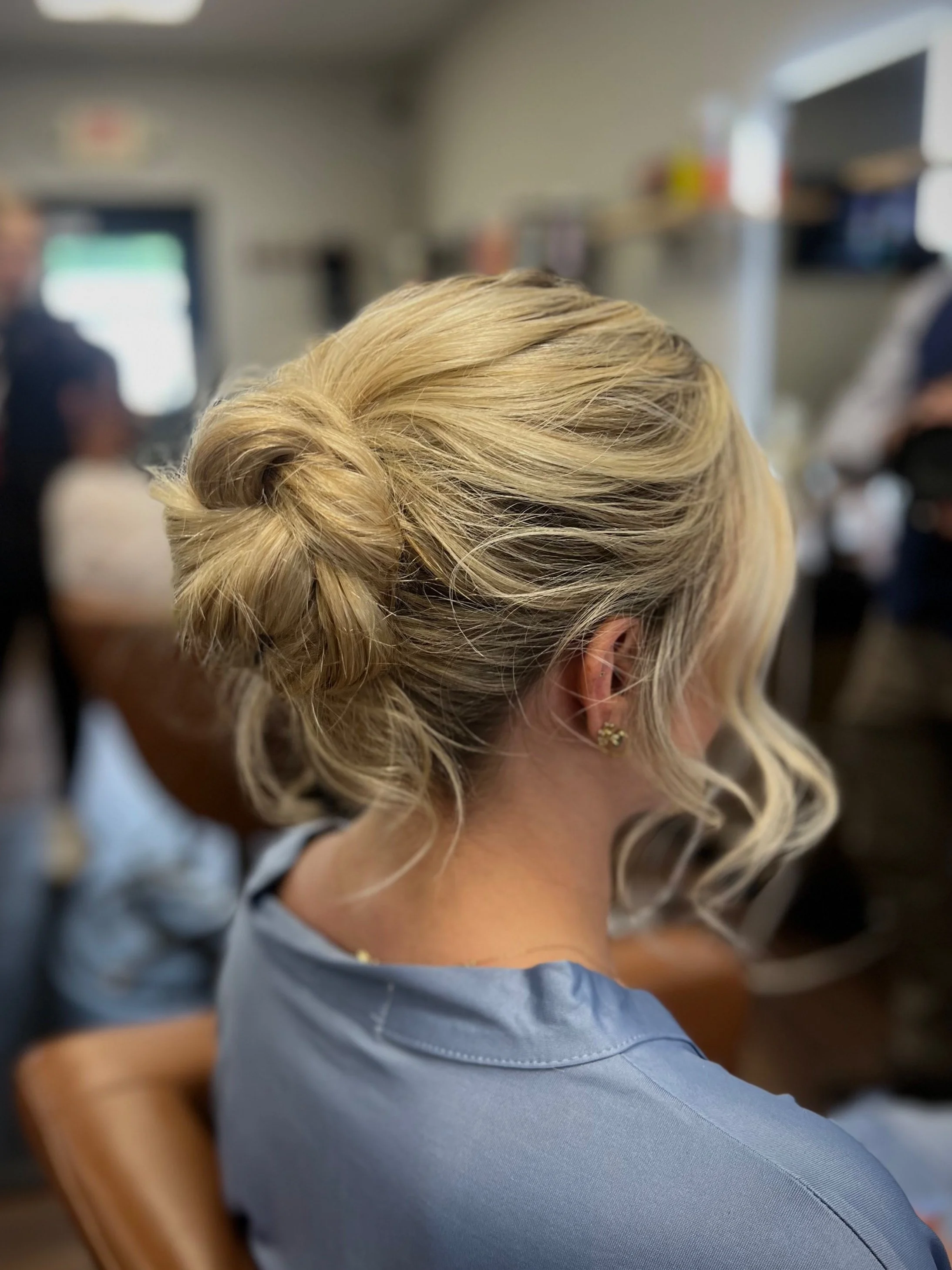 Blonde woman with her hair styled in an elegant low bun with loose curls framing her face, wearing pearl earrings and a light blue top, sitting in a busy indoor setting.