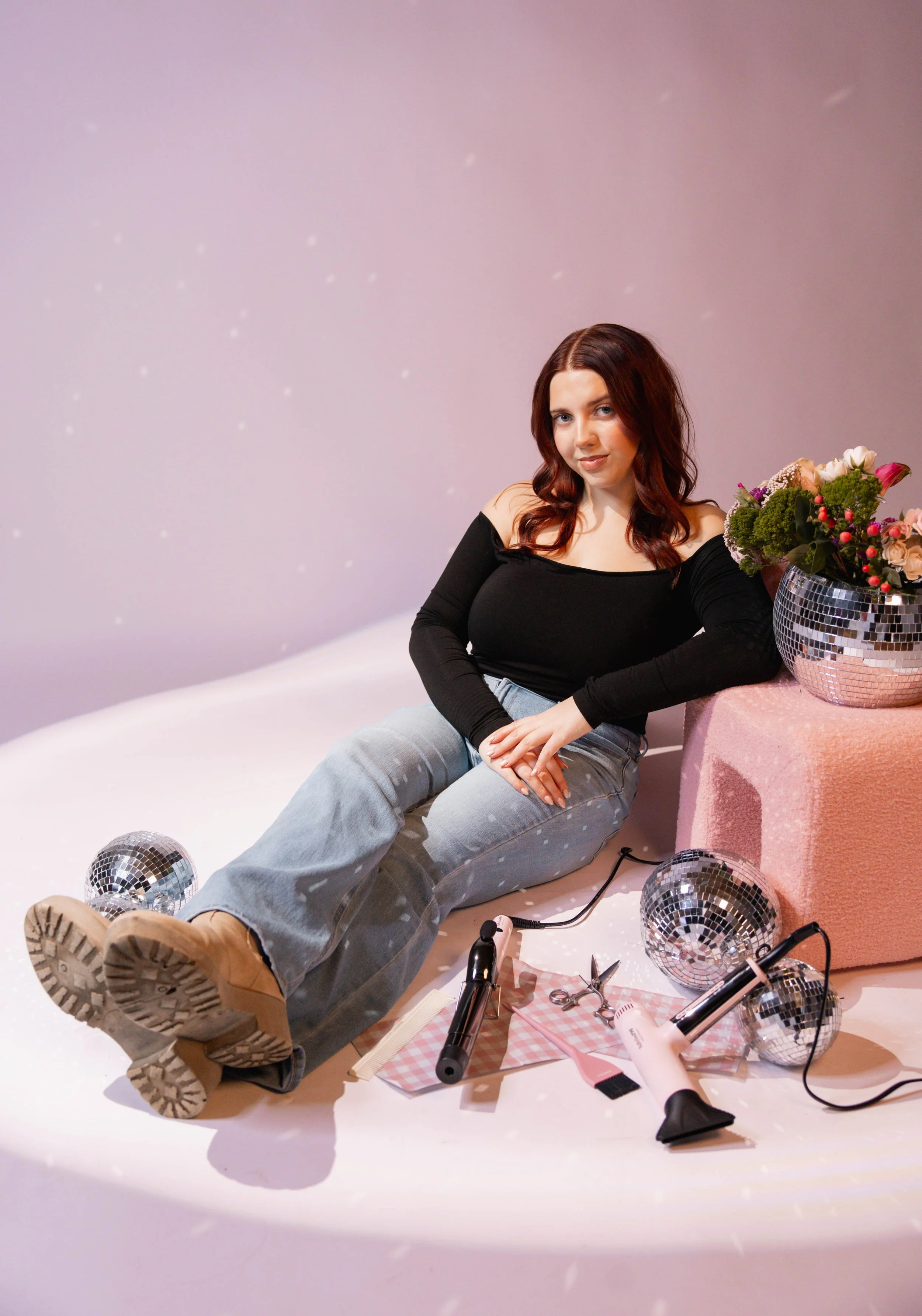 A person sitting on a bed using a silver laptop, holding a latte with latte art in their right hand, while their left hand is on the keyboard. The person is wearing light pink pants, and the bed has white bedding.
