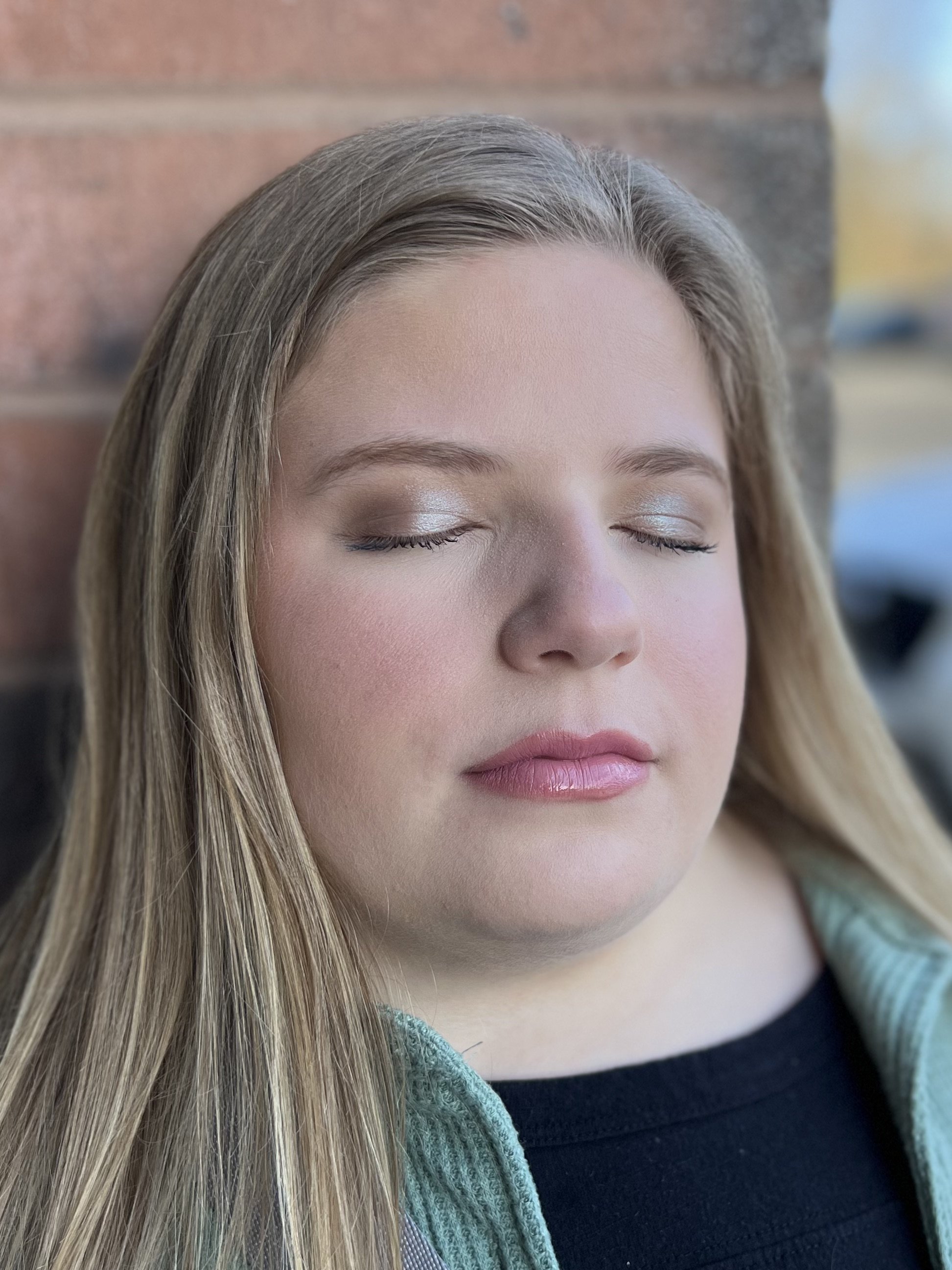 Close-up of a woman with closed eyes, wearing makeup, with long blonde hair, leaning against a brick wall.