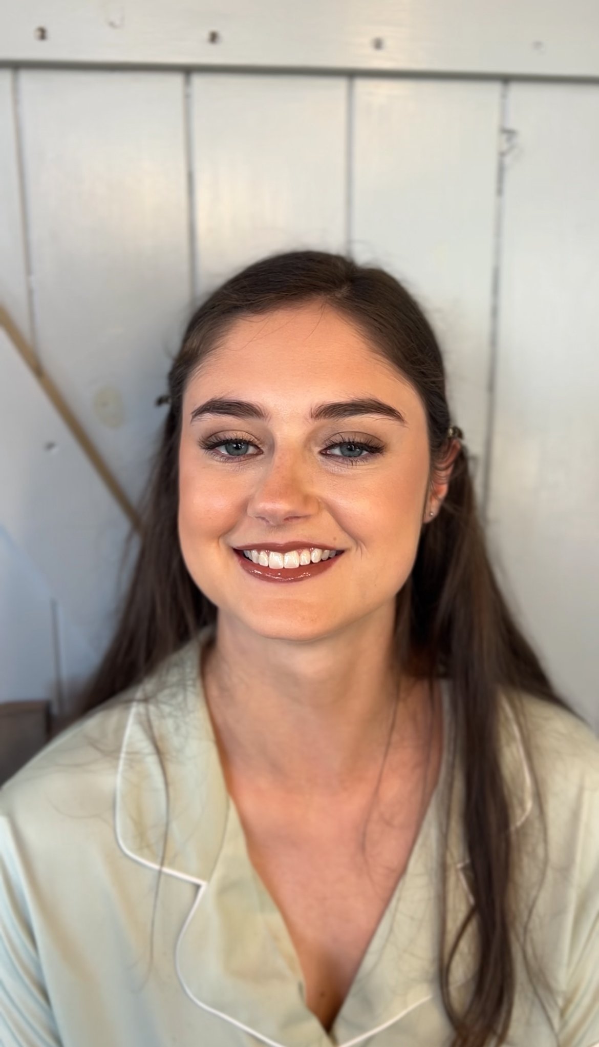 A young woman with long brown hair and blue eyes smiling, wearing light-colored pajamas, sitting in front of a white wooden wall.
