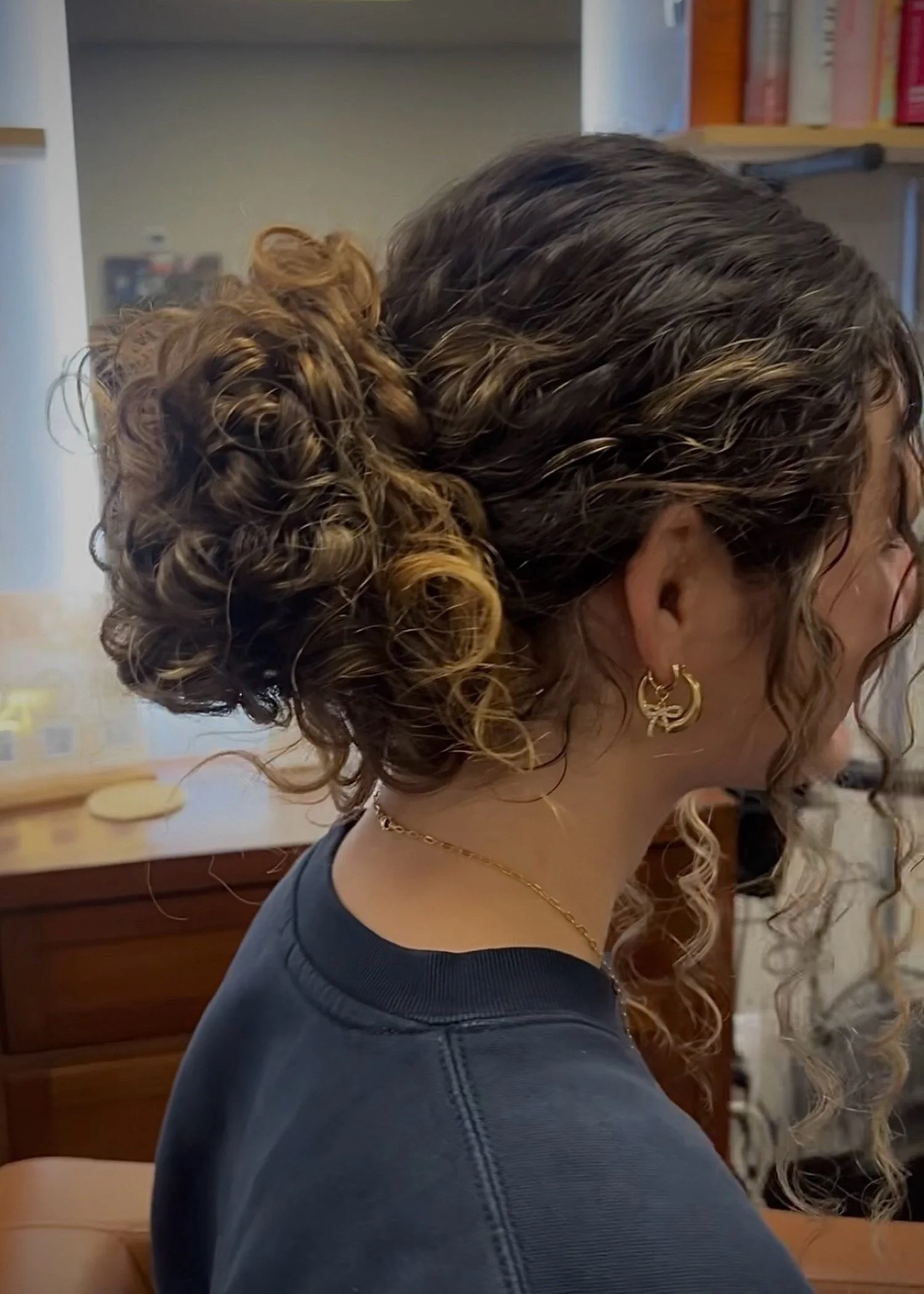 Side view of a woman with dark brown hair styled in an elegant updo with curls and highlights, wearing gold hoop earrings and a gold chain necklace, dressed in a dark shirt, in a room with wooden furniture and shelves in the background.