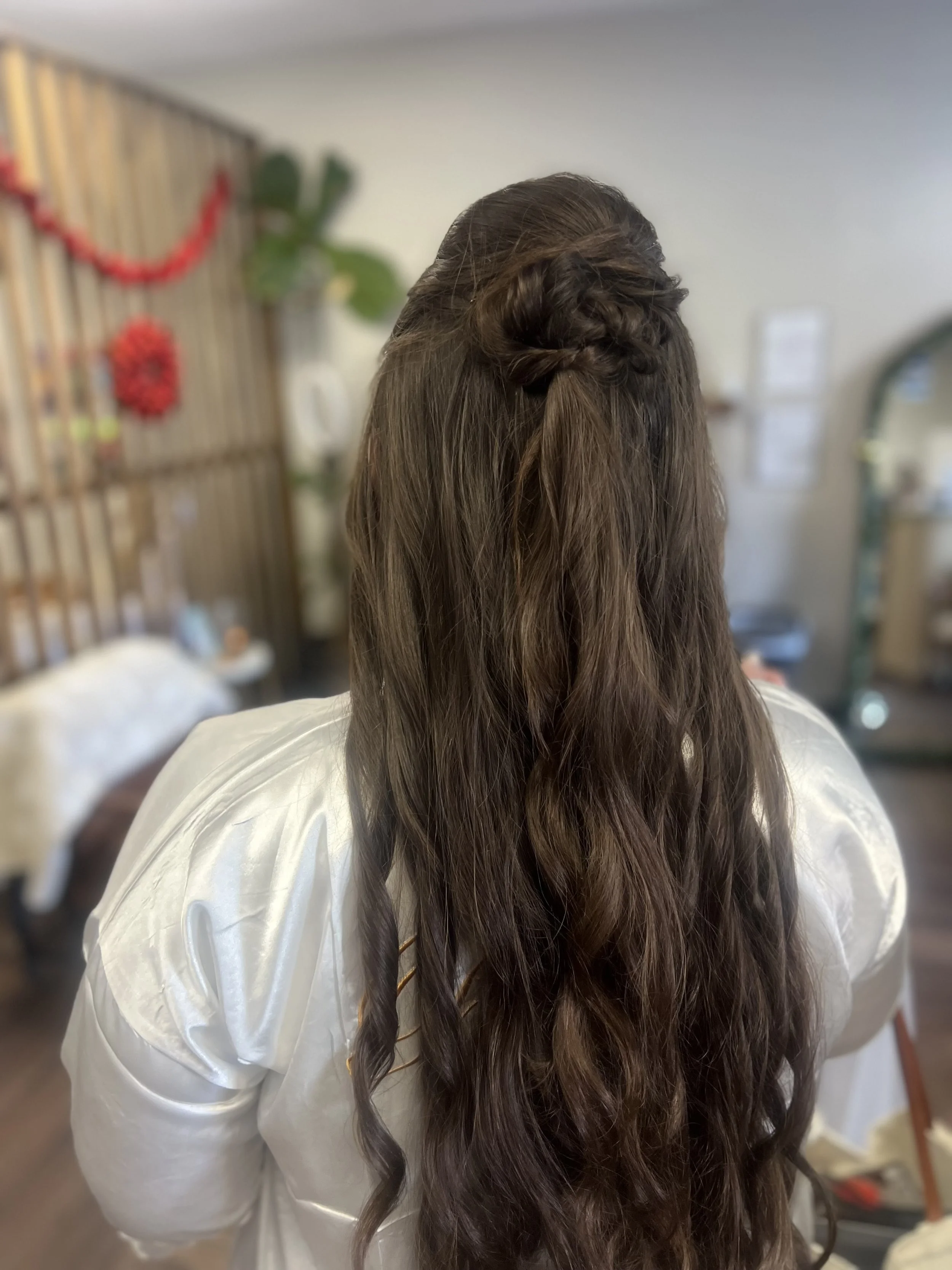 Back view of a woman with long, wavy brown hair styled with a half-up braid, wearing a satin white shirt, indoors with a decorated wall in the background.