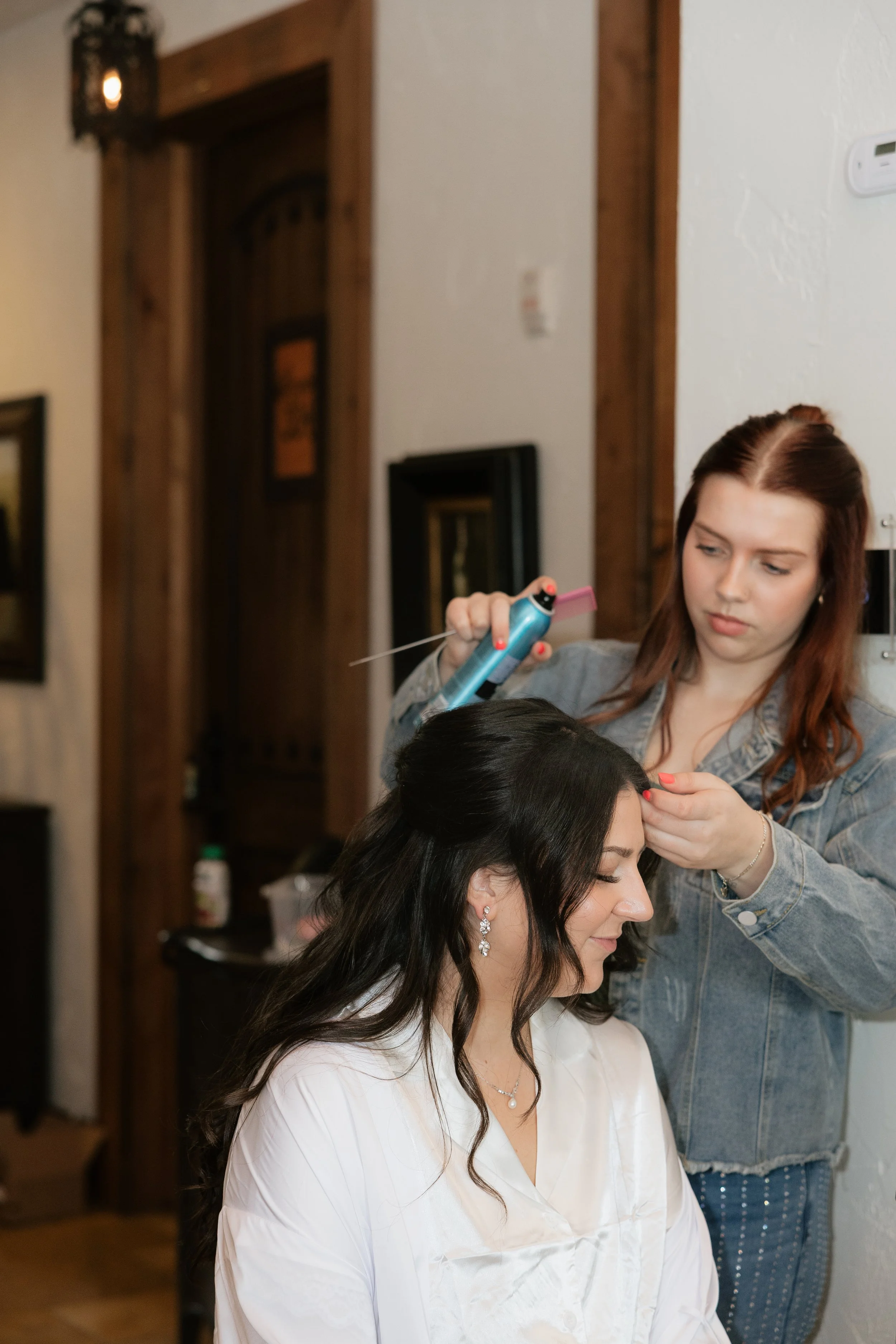 A woman with a satin robe sitting in a chair, getting her hair styled by a hairstylist in a black top. The woman is in a makeup room, with makeup and hairstyling tools on the counter, a glass of orange juice, and a large mirror reflecting her face.
