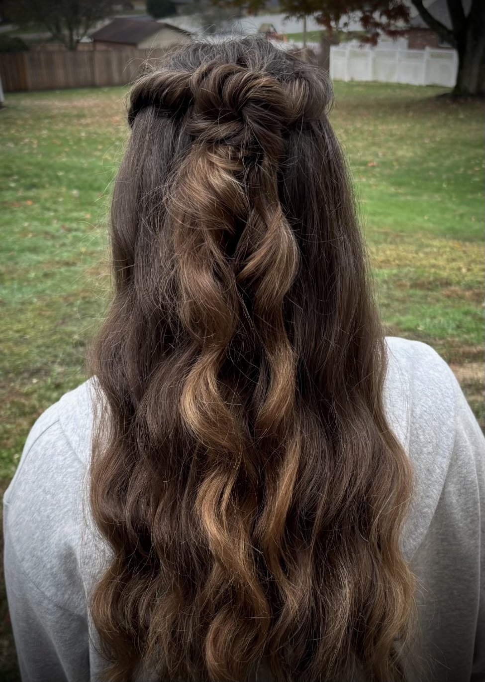 Back view of a woman with long wavy brown hair with lighter highlights, styled in loose curls with a small braid section at the top, outdoors in a yard with grass, trees, and houses in the background.