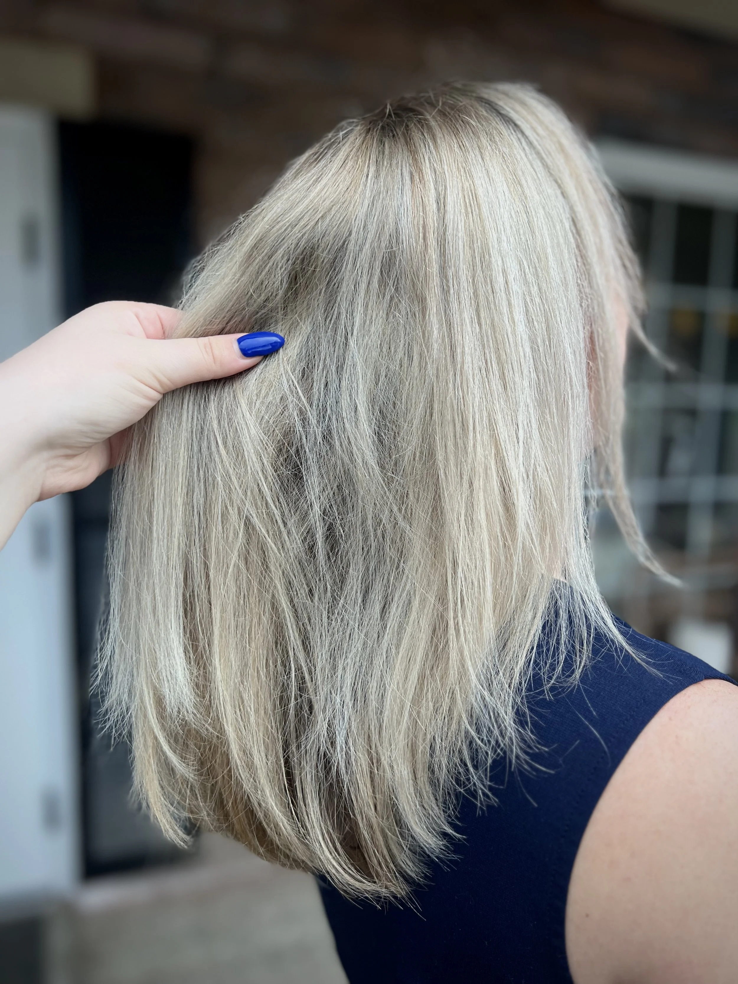 A person with blonde hair is holding a section of their hair, showing its texture and length. The background shows a blurred indoor environment.