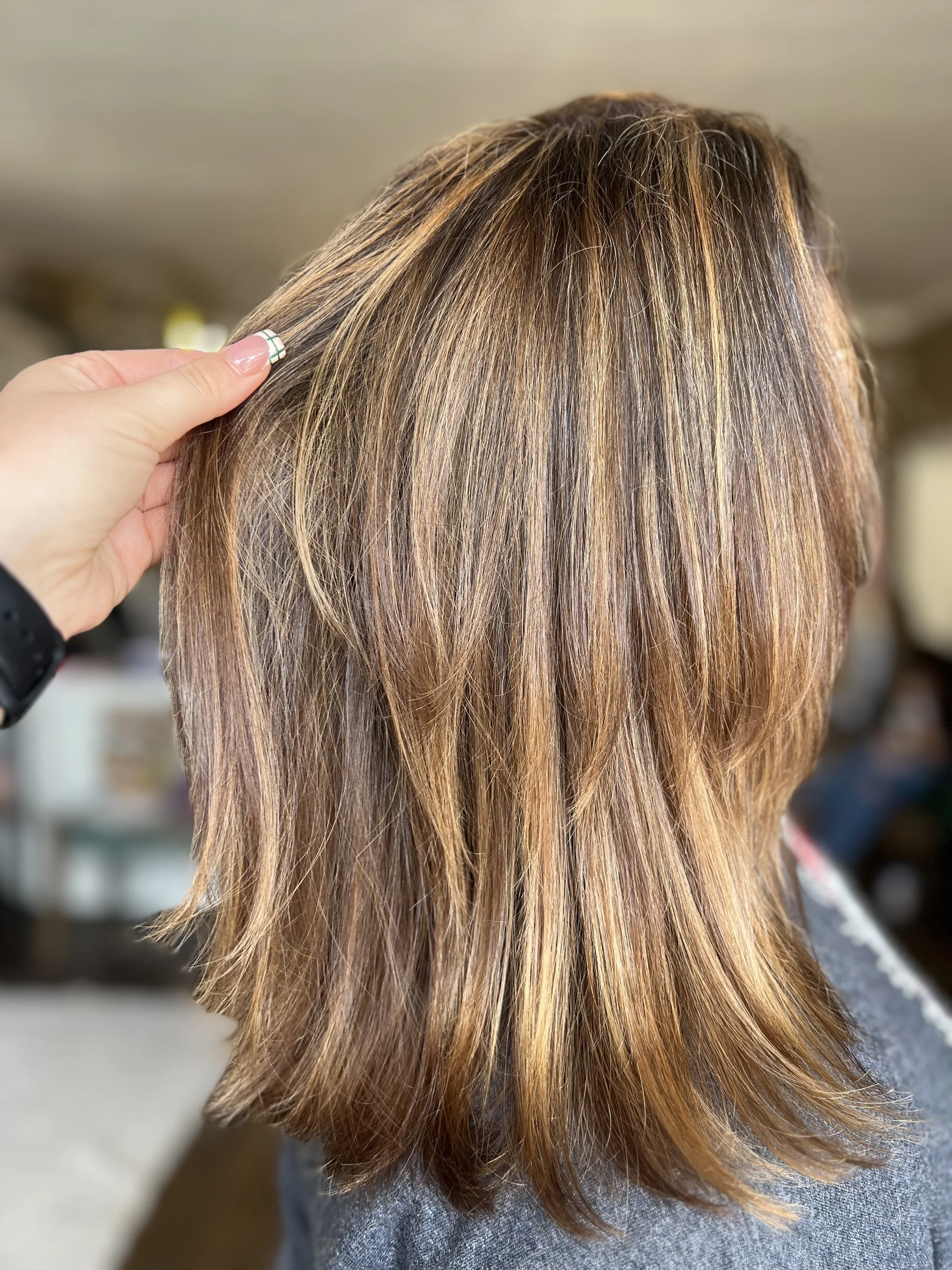 A person's shoulder-length hair with shades of brown, blonde, and light highlights, being styled or examined by a hand.