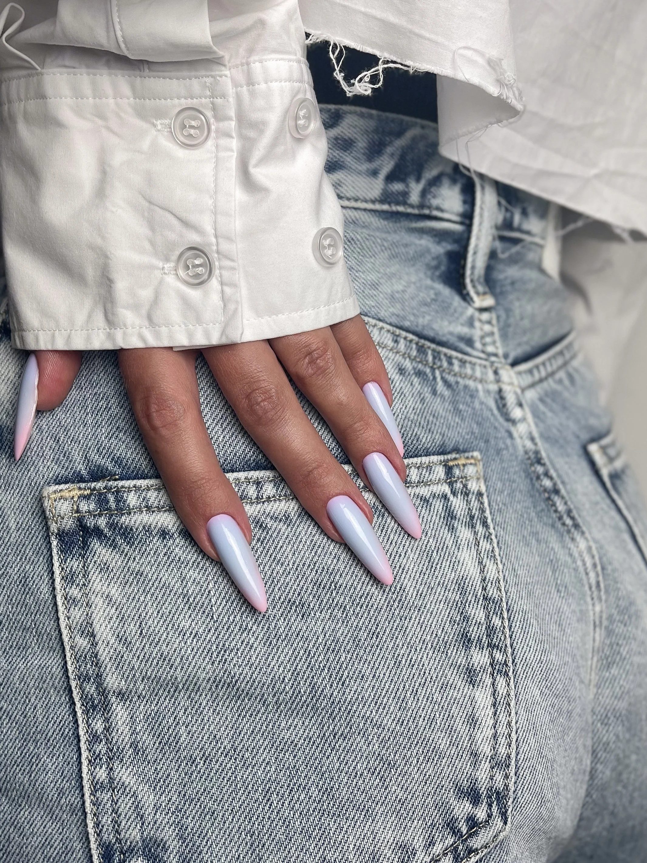 Close-up of a hand with long, glossy, light purple nails resting on the back pocket of light-wash denim jeans, with part of a white shirt and jacket visible.