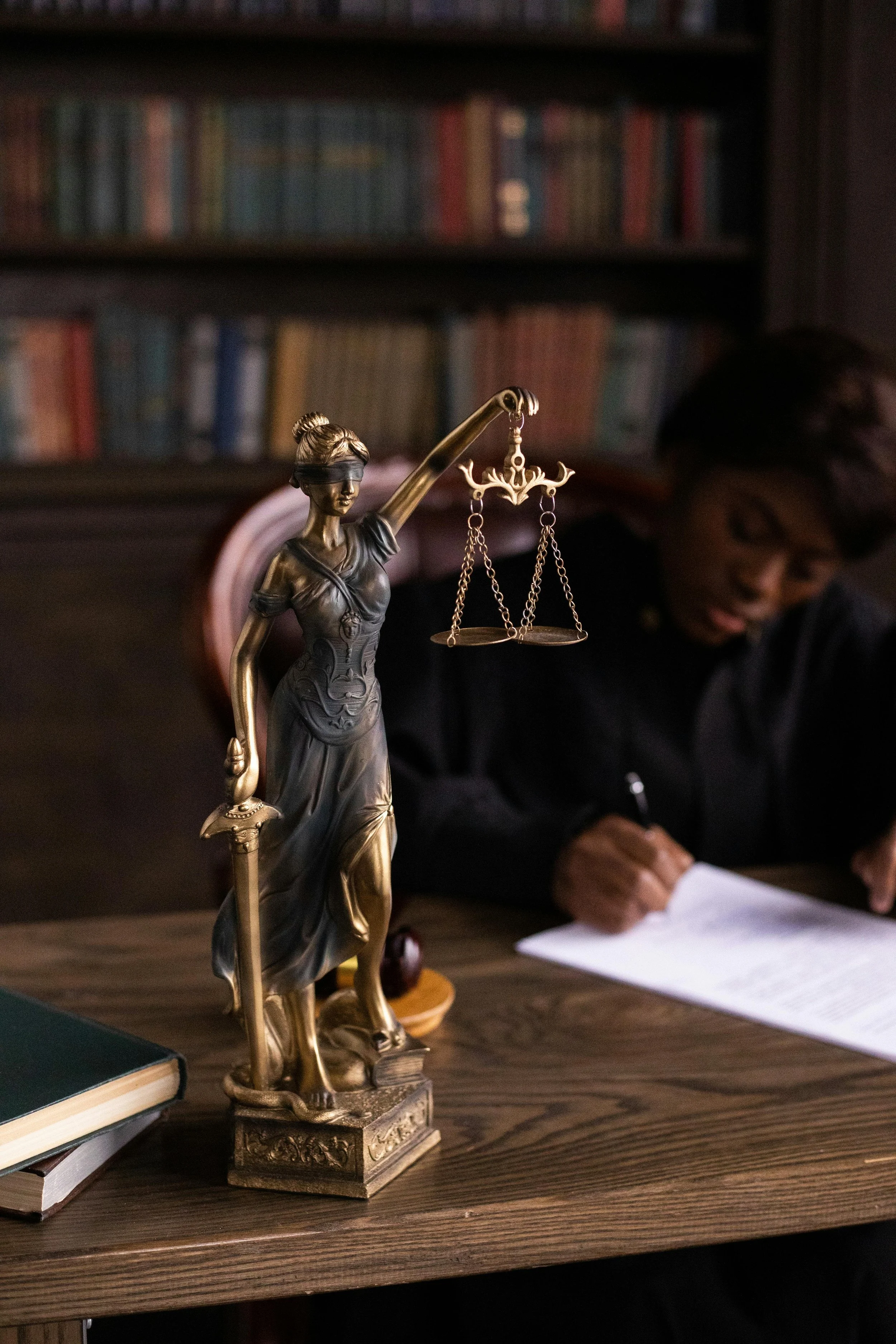 A bronze statue of Lady Justice with a blindfold and scales on a wooden desk in a law office or library, with a person writing at the desk in the background.