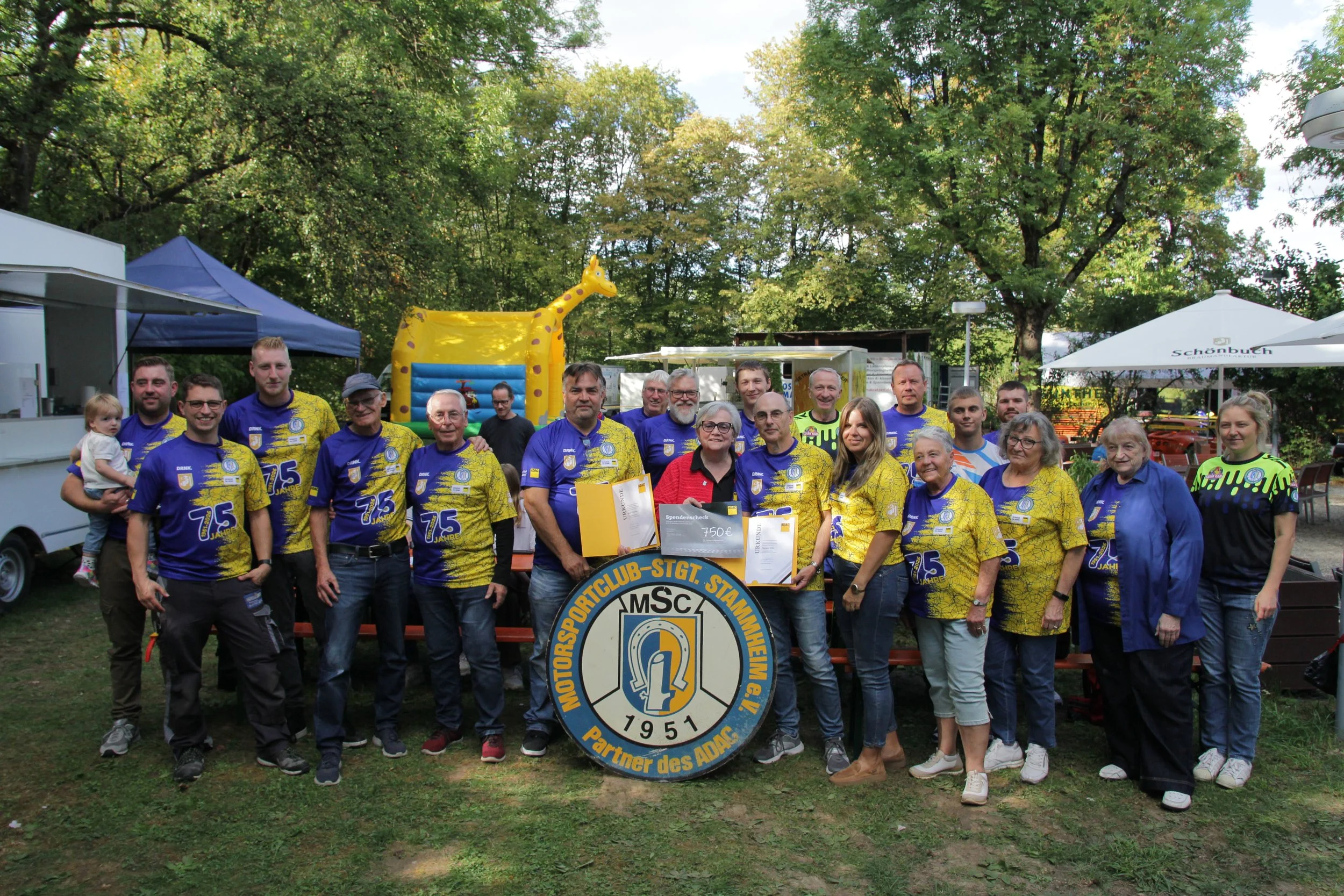 Gruppe von Menschen, die bei einer Feier im Freien zusammenstehen, einige in blauen Trikots mit 75 Jahren Jubiläum, im Hintergrund ein gelbes Giraffen-Ballon und ein Vereinswappen, umgeben von Bäumen.