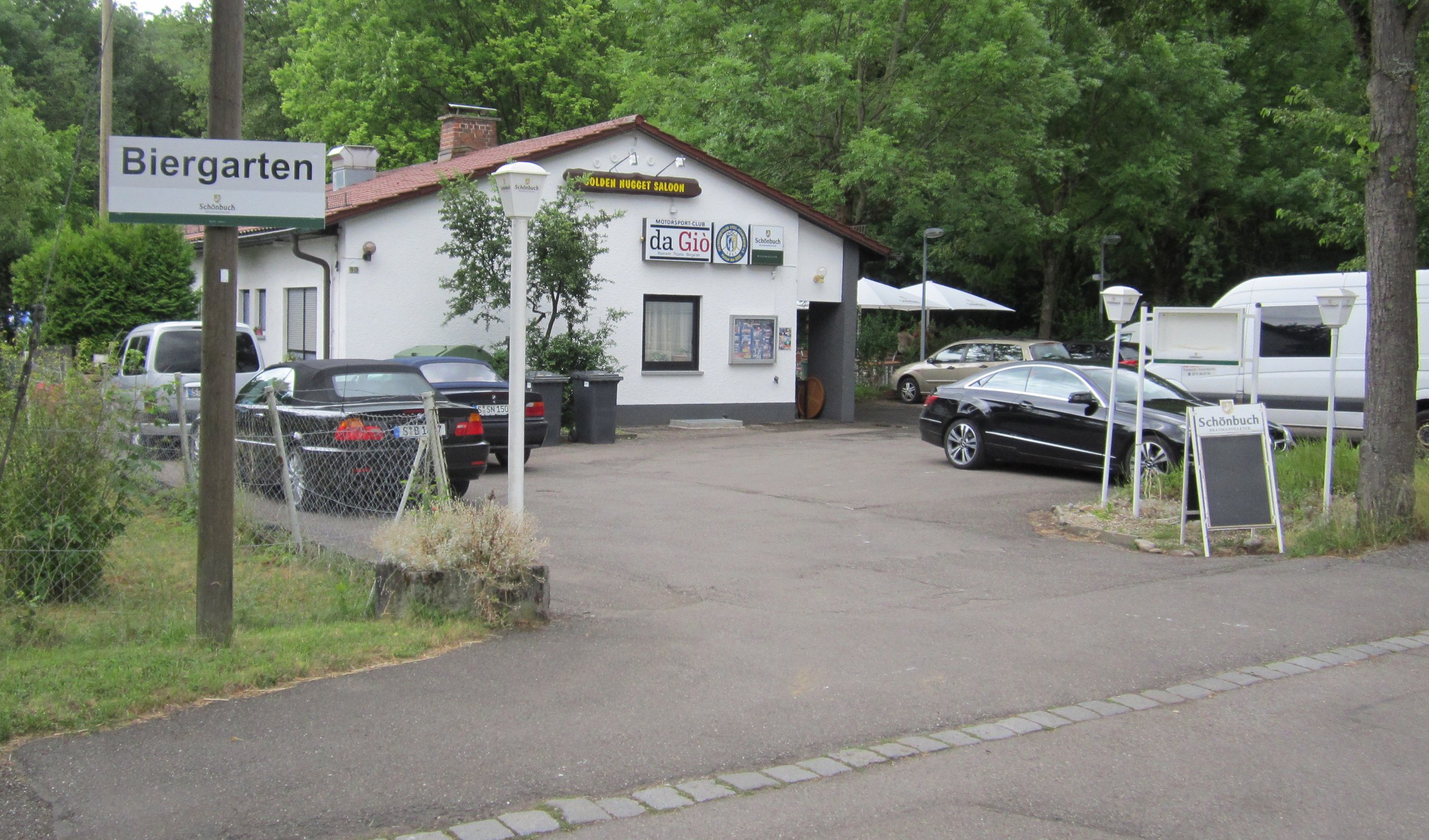 Ein Parkplatz vor einem kleinen Gebäude mit mehreren Autos und einem großen Baum im Hintergrund. Es befindet sich ein Schild mit der Aufschrift 'Biergarten' und weitere Schilder am Gebäude.