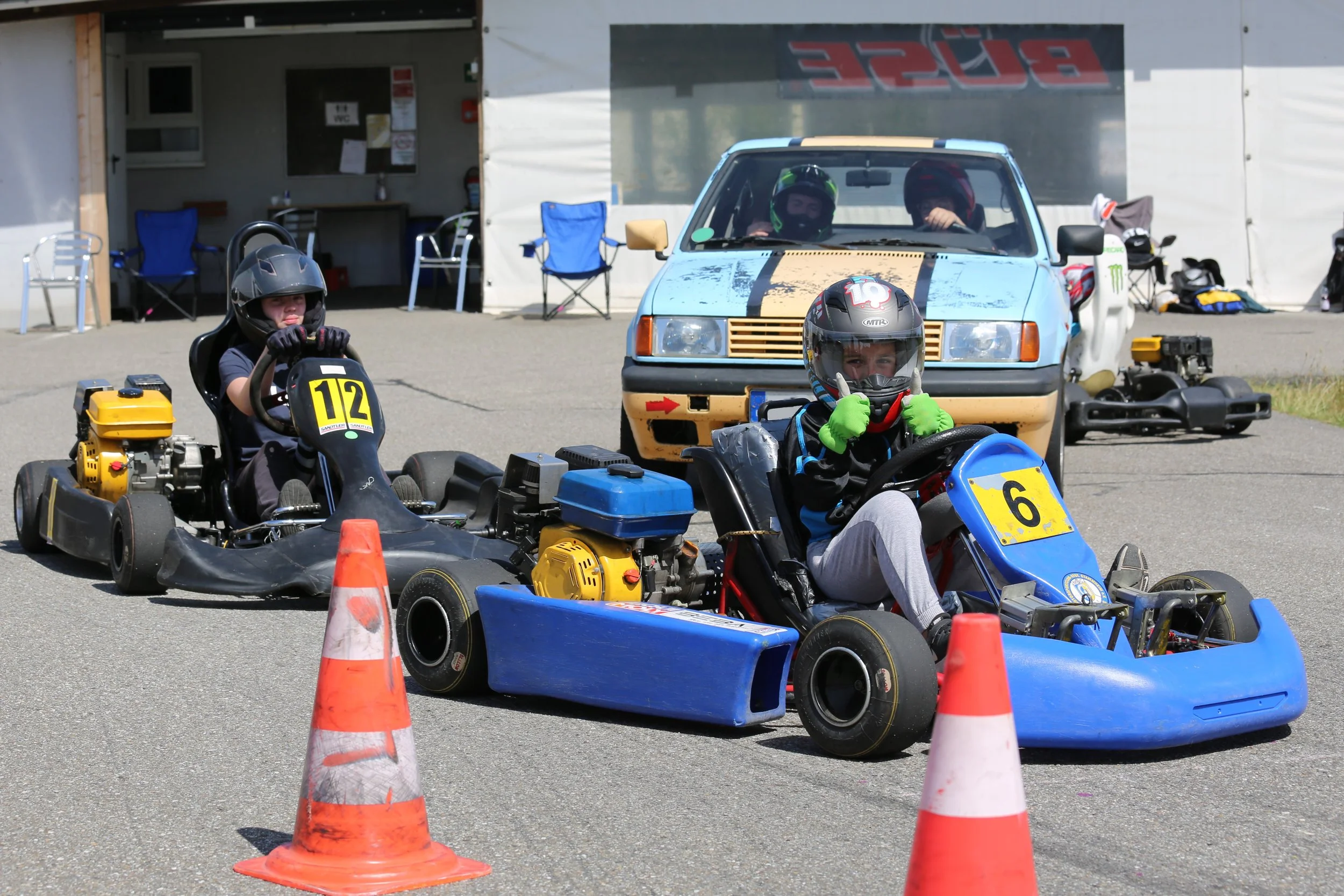 Zwei Kinder fahren mit Go-Karts zwischen orangenen Verkehrsampeln auf einem Parkplatz. Beide tragen Helme und zeigen lautstark Daumen nach oben.