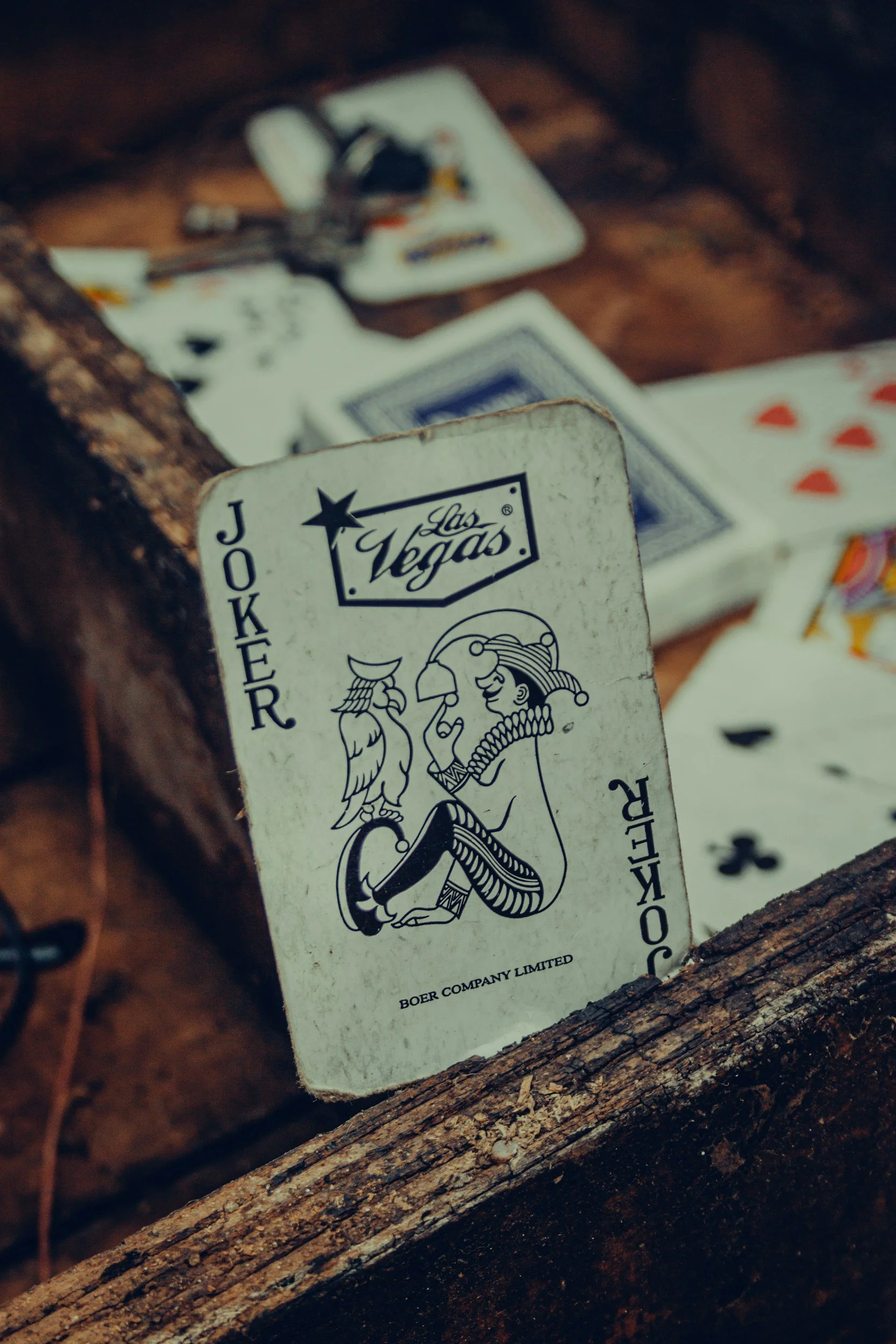 A deck of cards with a joker card featuring a stylized figure of a woman with feathers and jewelry, leaning against a wooden surface with other playing cards spread out in the background.