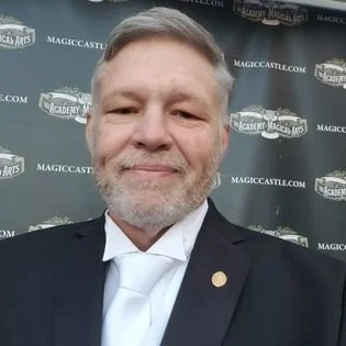 A man with gray hair and beard wearing a dark suit and white shirt, taking a selfie in front of a backdrop with 'Magic Castle' logos.