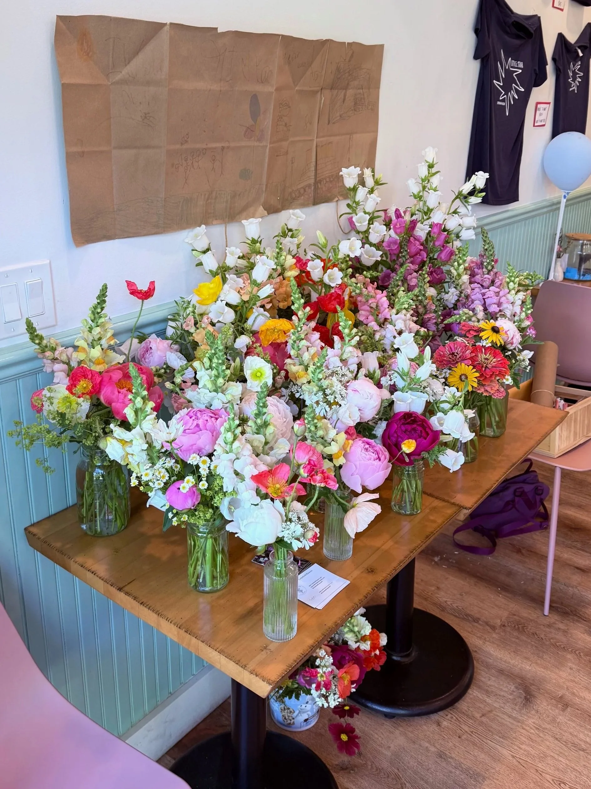 A table displays multiple glass vases filled with colorful flowers, including white, pink, yellow, and red blooms, in a room with a white wall, a brown paper art piece, and black T-shirts hanging on the wall.