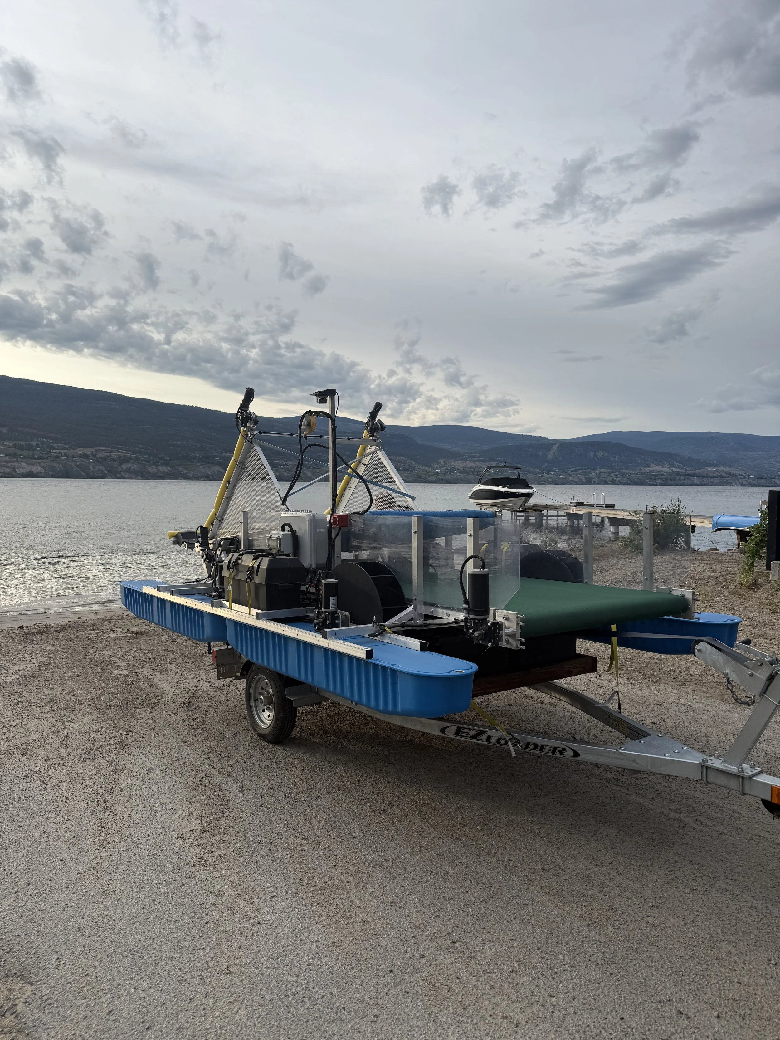 A mechanized personal watercraft on a trailer parked on a gravel surface near a body of water with hills and a cloudy sky in the background.