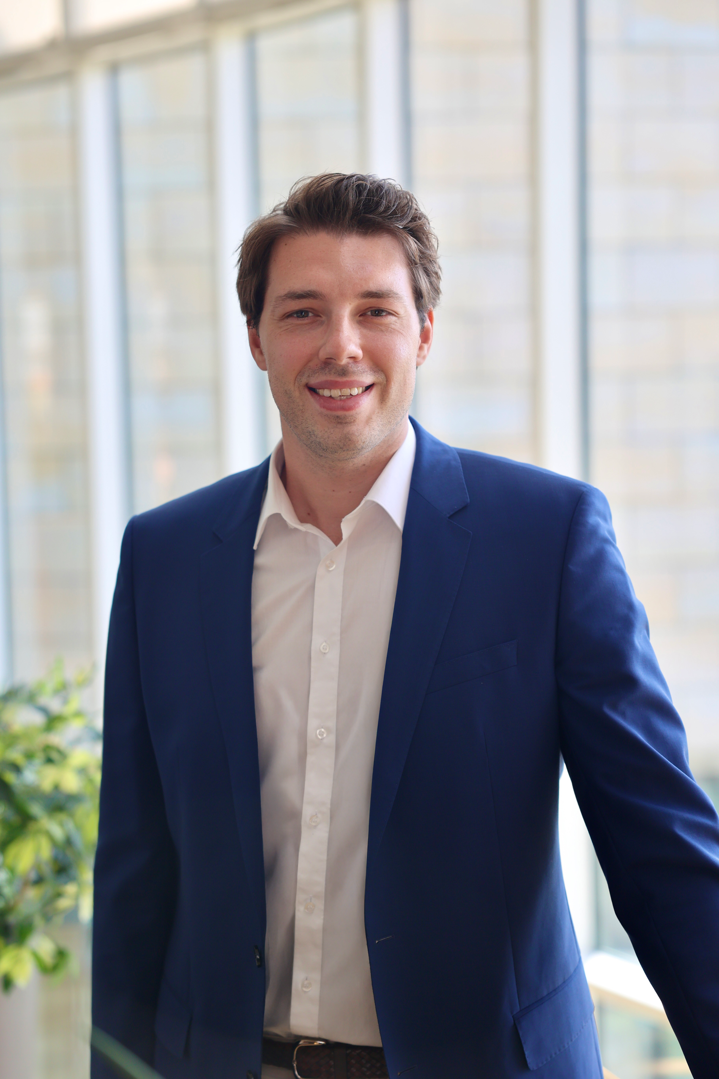 Yannick Breitsch in einem blauen Anzug mit weißem Hemd steht in einem hellen Gebäude mit großen Fenstern, lächelnd in die Kamera.