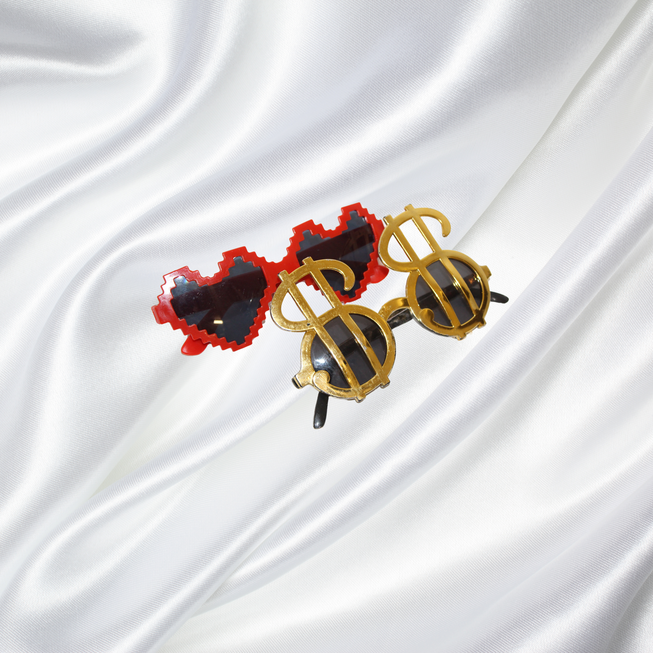Pair of novelty sunglasses, one with pixelated red heart-shaped frames and dark lenses, the other with gold dollar sign frames and dark lenses, resting on a shiny white satin fabric.