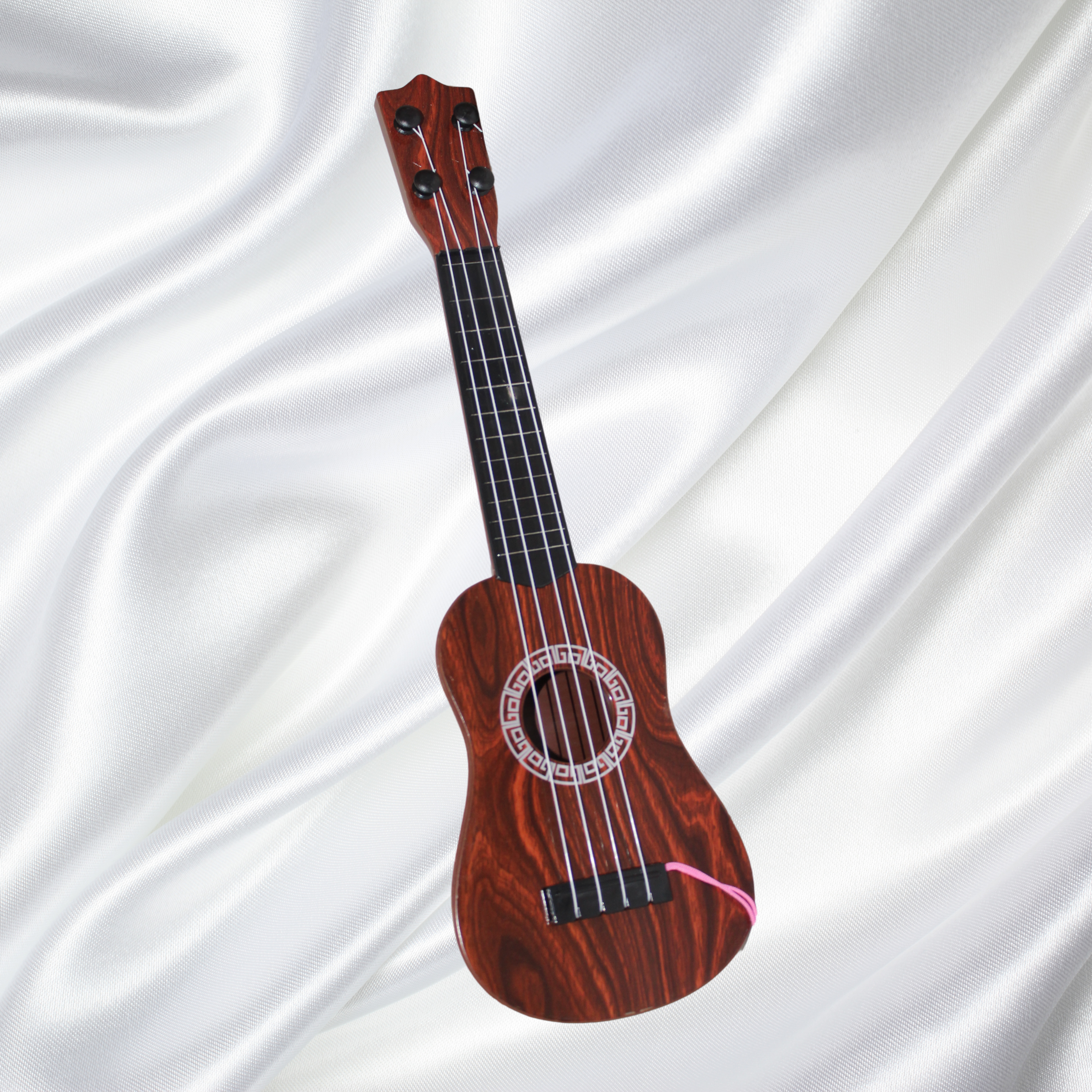 A wooden ukulele with black tuning pegs and four strings, resting on white satin fabric.