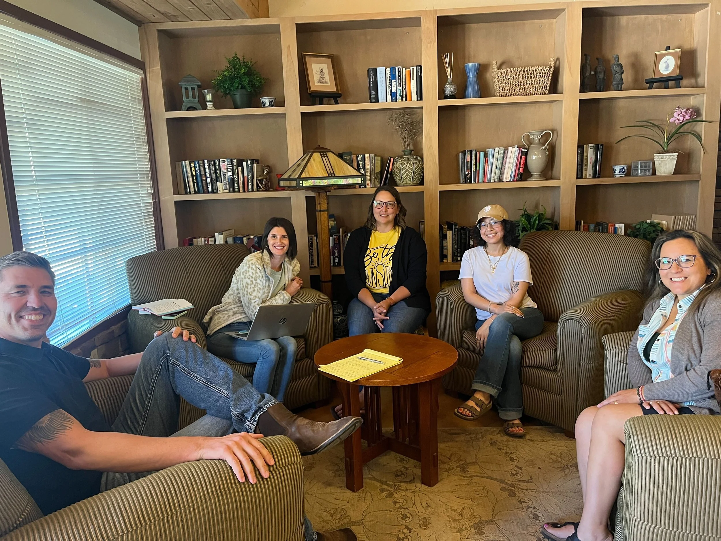 Five people sitting in a cozy living room with bookshelves, engaging in a casual meeting or conversation.