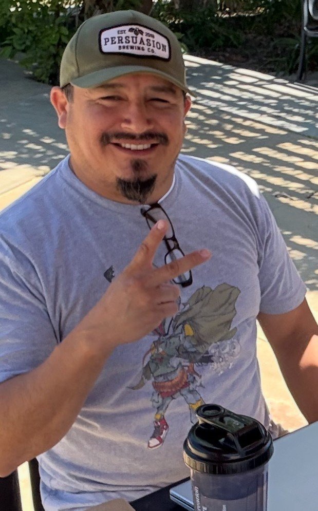 A smiling man wearing a baseball cap with the logo "Persuasion Brewing Co." and a T-shirt featuring a cartoon character, sitting outdoors at a table with a coffee cup and glasses.