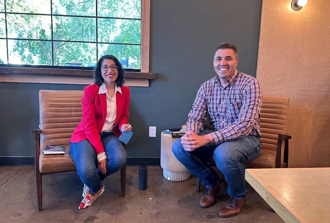 Two smiling people sitting on separate chairs in a room with a large window showing trees outside. The woman on the left is wearing a red blazer, white shirt, and jeans, holding a phone and sitting with legs crossed. The man on the right is wearing a plaid shirt and jeans, sitting with hands clasped. There is a small side table between them with a cup and other items, and a textured ottoman beneath the window.