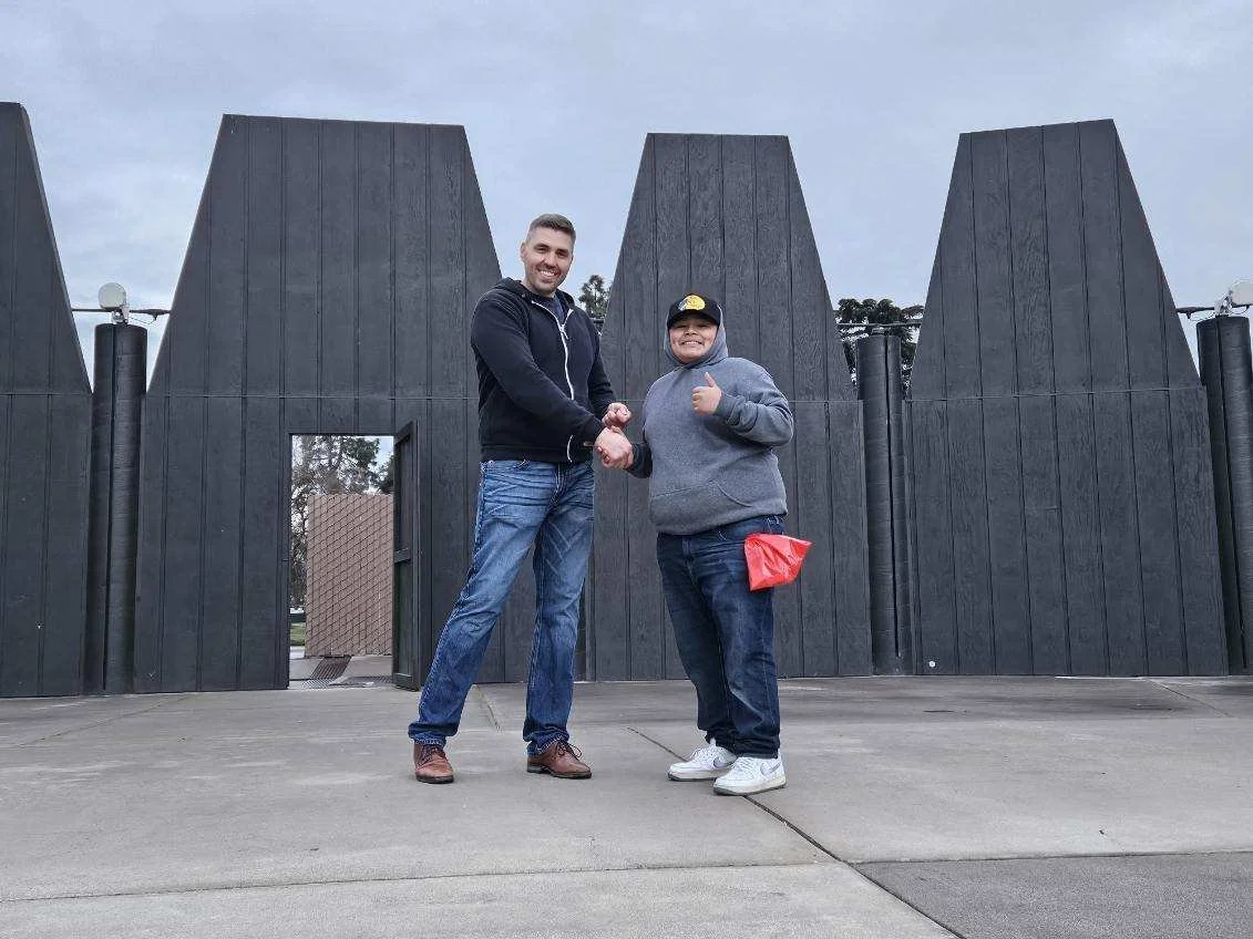 Two men shaking hands in front of a large, black, metallic structure with pointed peaks, on a cloudy day.