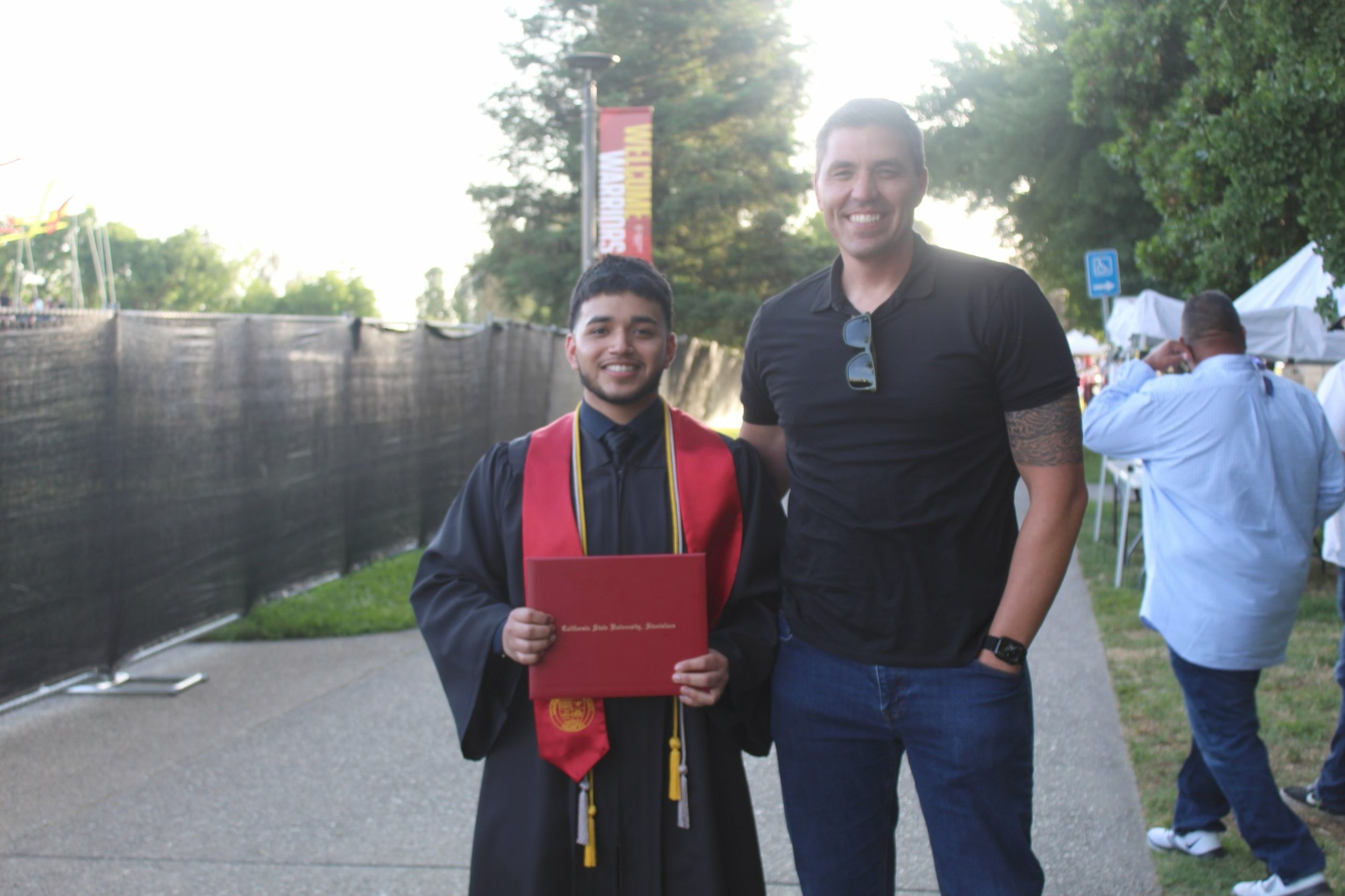 A young man in a graduation gown holding diploma, standing next to an adult man, both smiling outdoors during a graduation celebration.